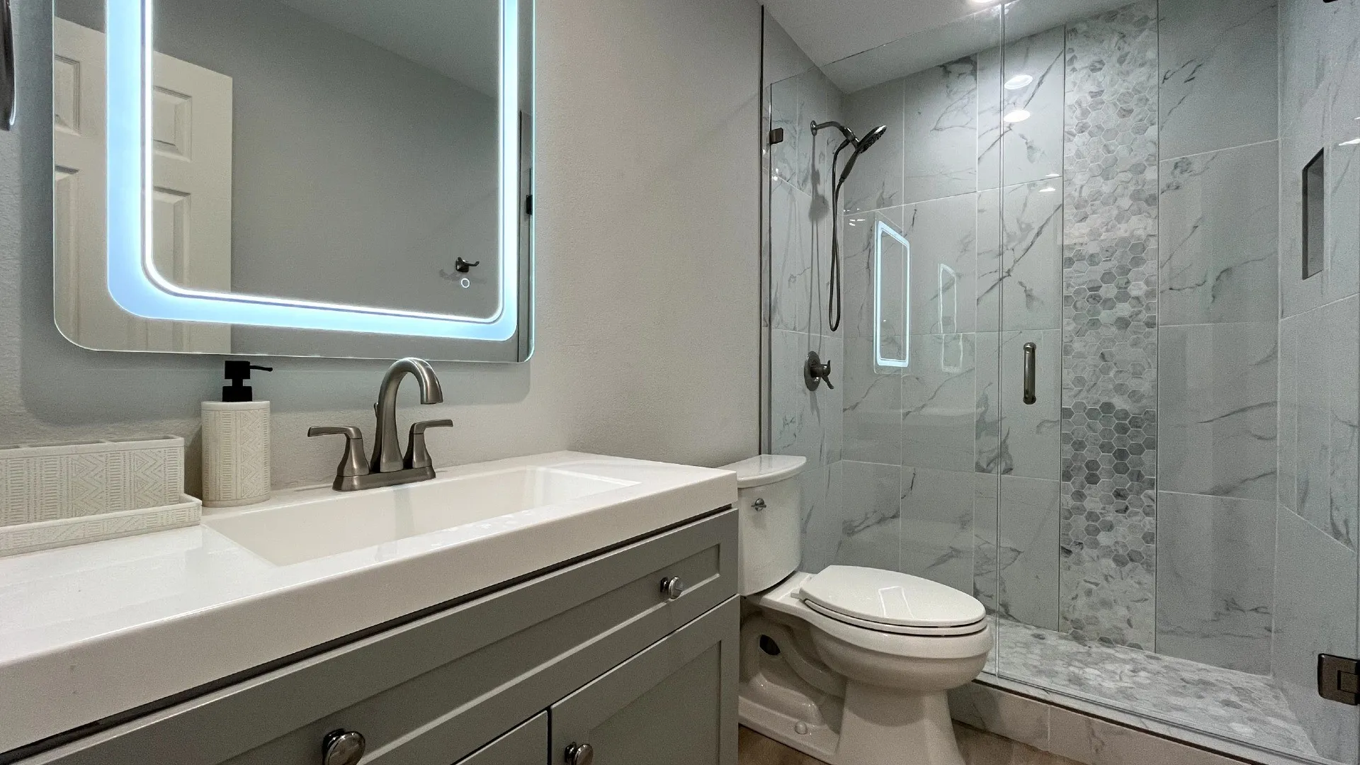 Vanity with integrated sink, illuminated mirror, glass-enclosed shower, wall-mounted showerhead, and tiled shower surround with mosaic accent