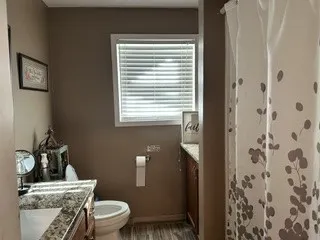 Wood-finish flooring with a white toilet, a window with horizontal blinds, and a vanity featuring a patterned countertop and wood cabinetry