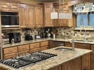 Wood cabinetry, stone countertops, stone tile backsplash, stainless steel appliances, and a kitchen island with cooktop and sink