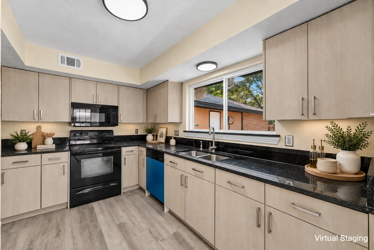 Modern kitchen featuring light wood-finish cabinetry with chrome hardware, black quartz countertops, black stainless steel appliances including a built-in wall oven and range, stainless steel sink with chrome faucet, wood-finish flooring, and abundant natural light from large windows overlooking the exterior