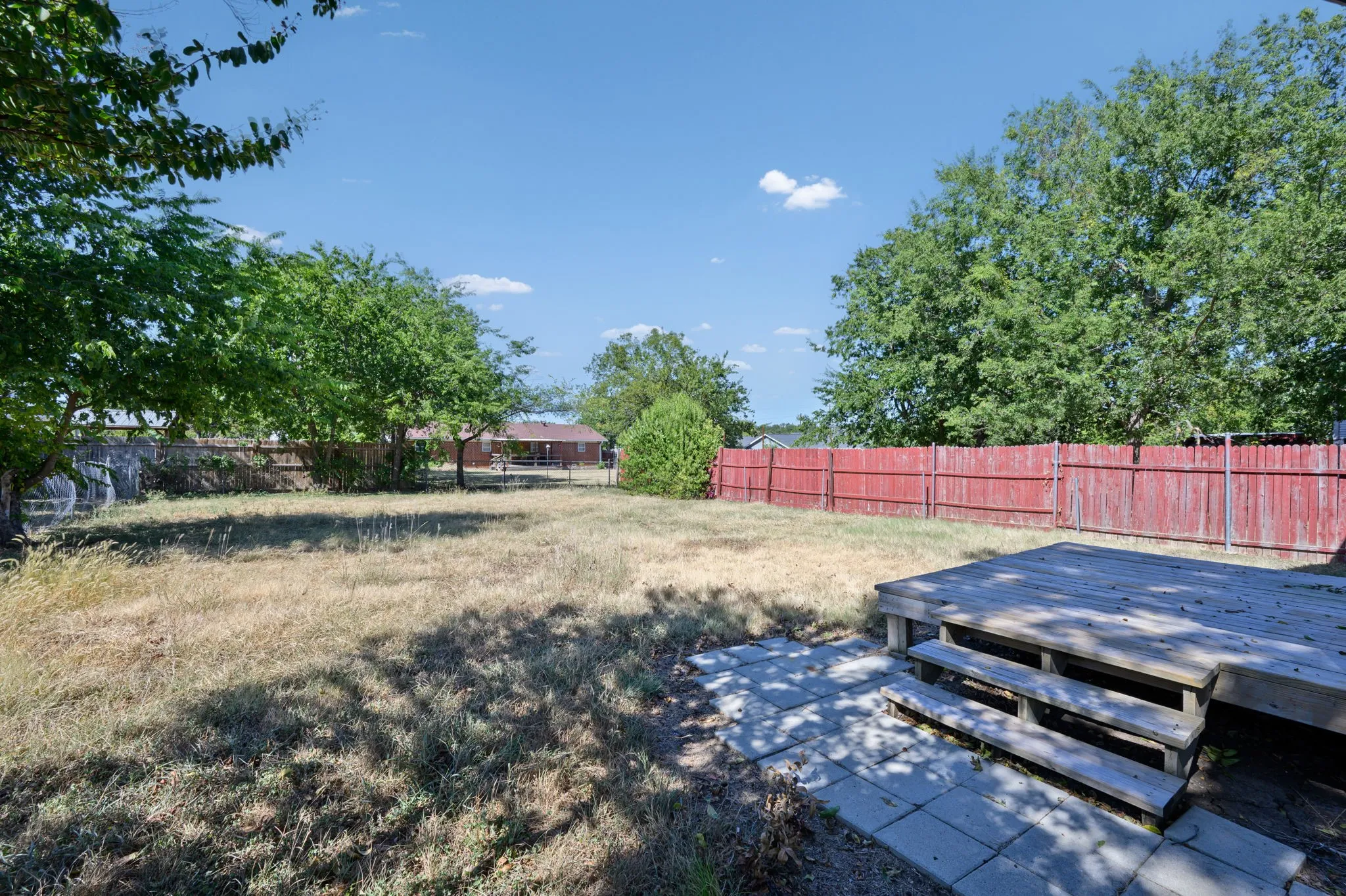 Expansive backyard featuring a wood deck with steps, red-stained privacy fencing, and mature trees