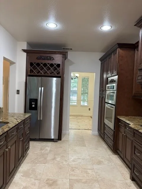 Kitchen featuring recessed lighting, extensive dark wood cabinetry, light-toned stone-finish flooring, granite countertops, and built-in stainless steel appliances including a refrigerator and double oven
