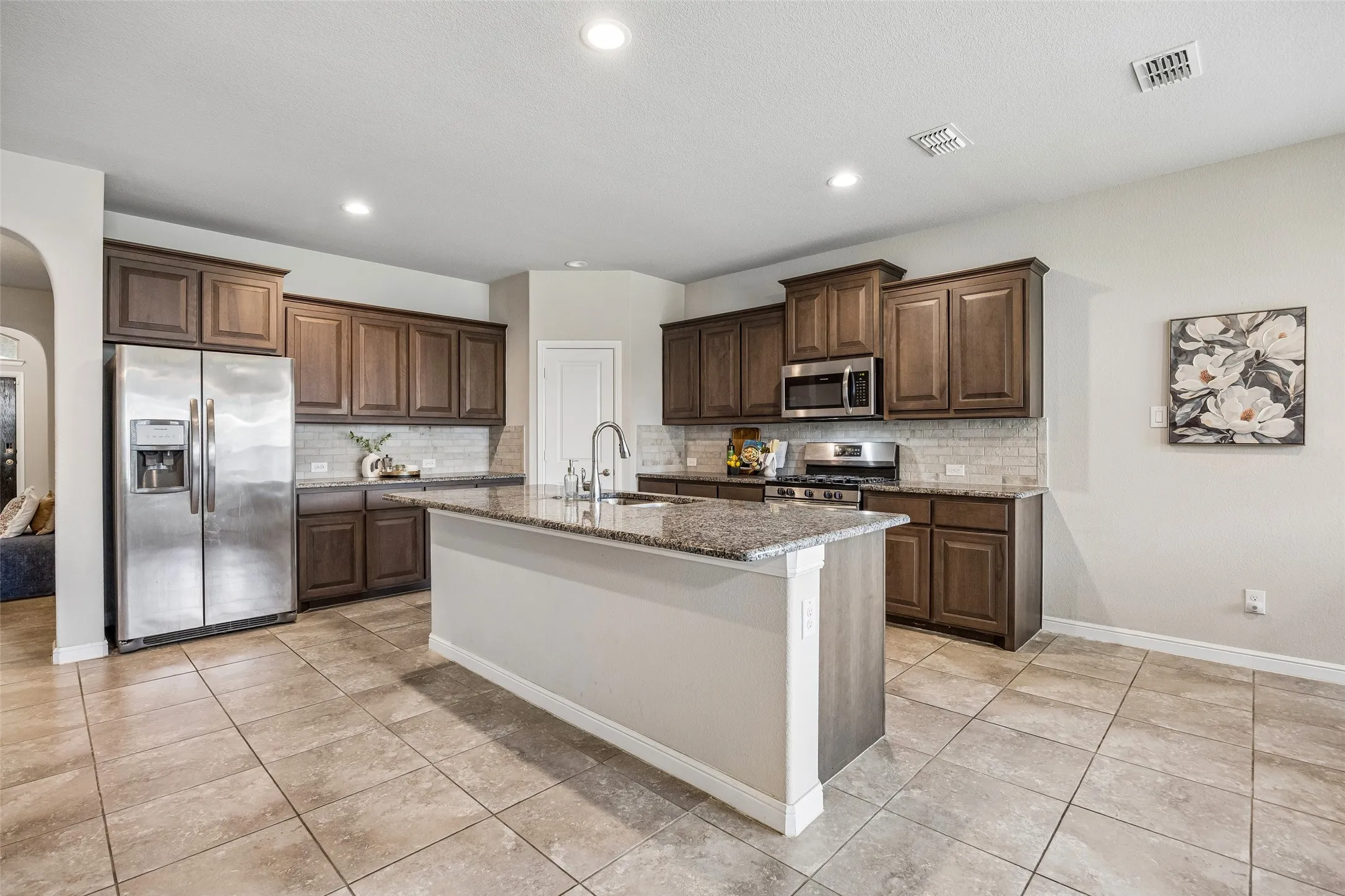 Spacious kitchen featuring rich wood cabinetry, stainless steel appliances including a side-by-side refrigerator, granite countertops, a large center island with white base and seating area, recessed lighting, and tile flooring throughout the open layout