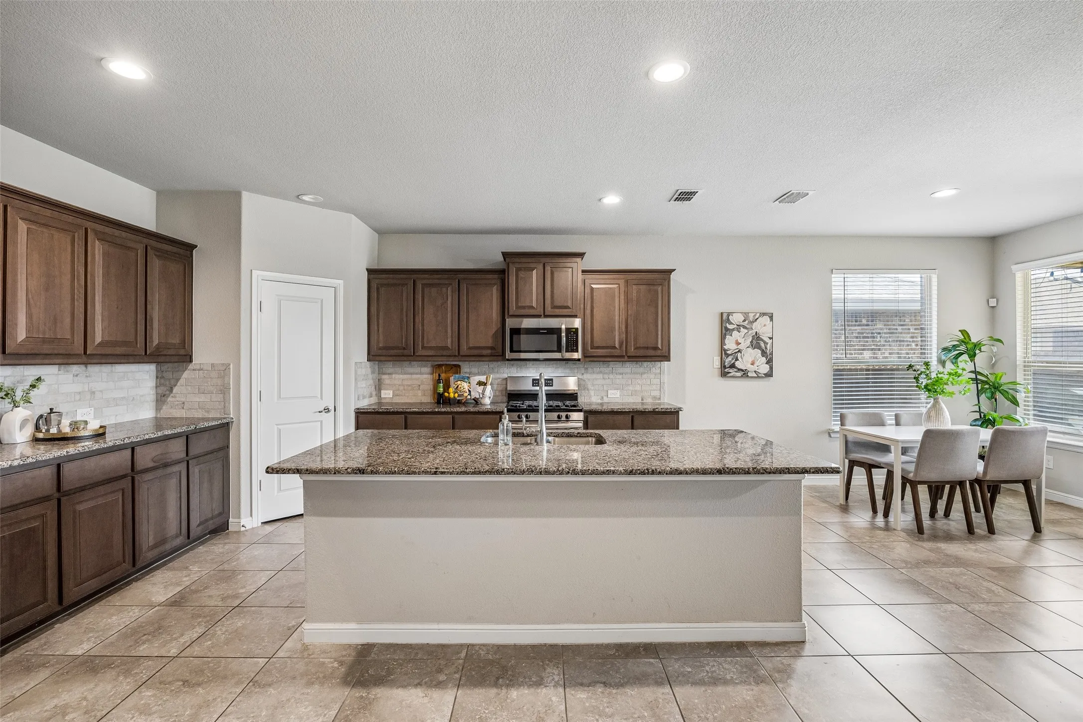 Spacious open-concept kitchen and dining area featuring rich wood cabinetry, stainless steel appliances, granite countertops, a large center island with seating, and tile flooring throughout