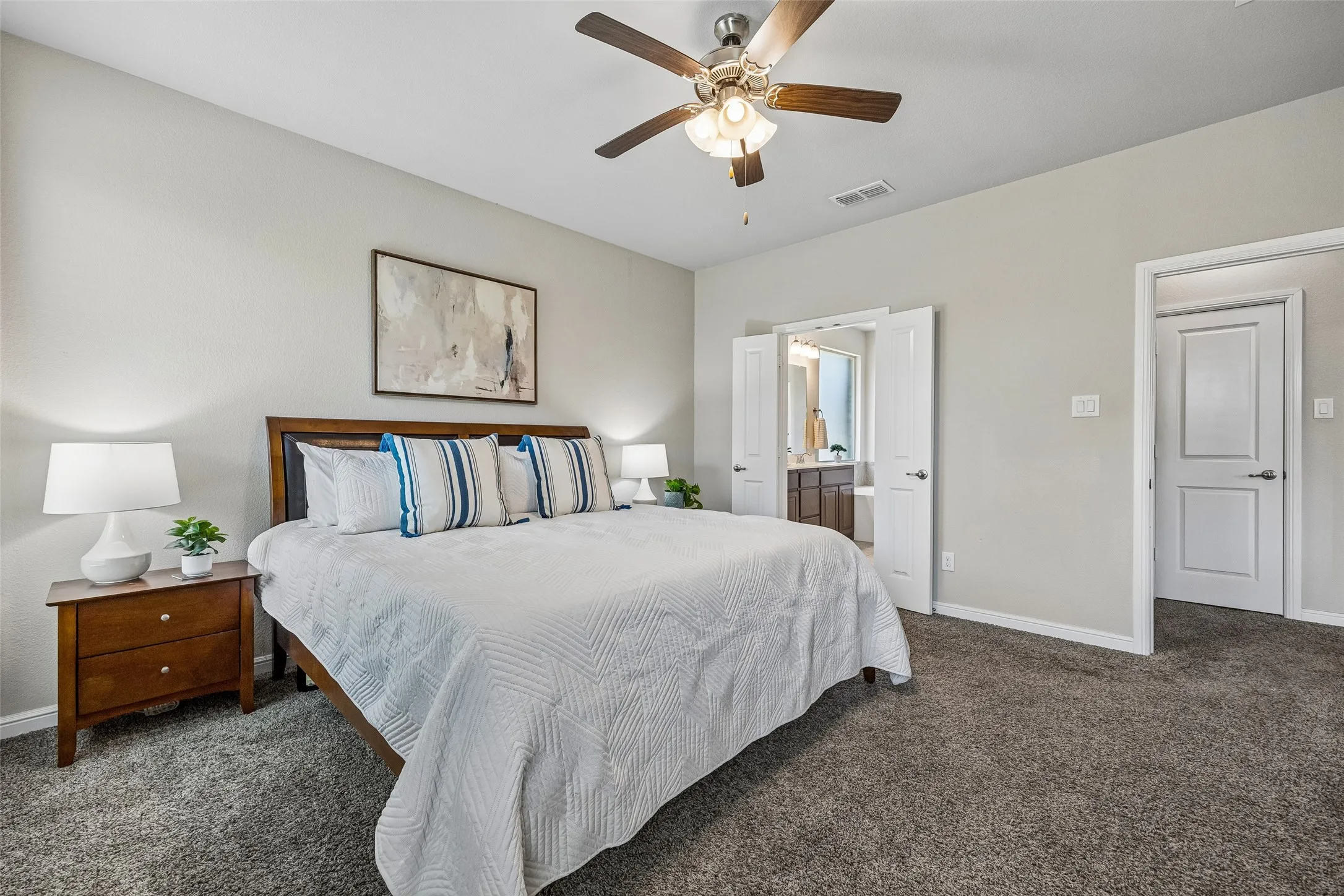 Spacious primary bedroom with wood-frame bed, soft-close white panel doors, ceiling fan with integrated lighting, neutral wall finishes, and carpeted flooring
