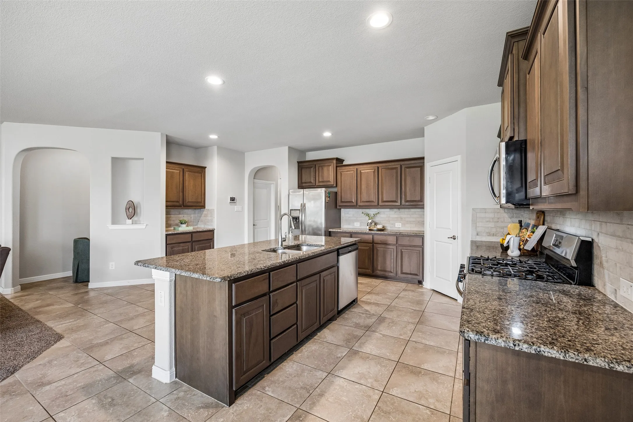 Kitchen featuring a central island with an undermount sink, granite countertops, wood cabinetry, stainless steel appliances, and tile flooring