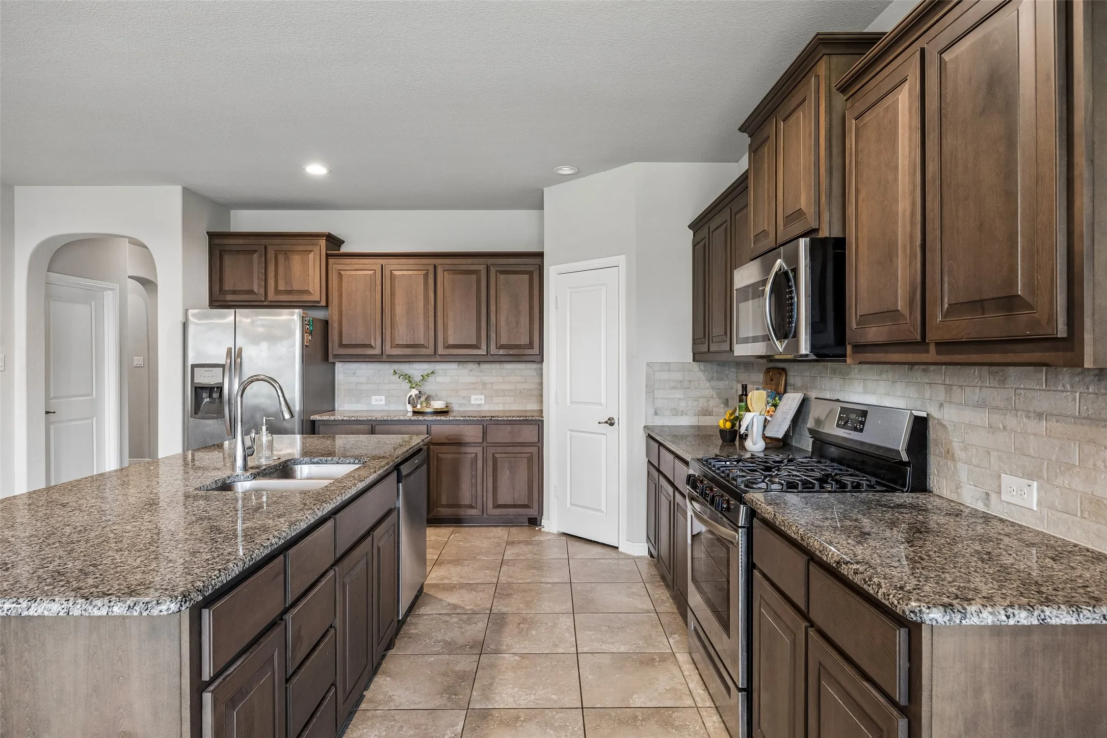 Spacious kitchen with granite countertops, stainless steel appliances, dark wood cabinetry, tile flooring, and a subway tile backsplash