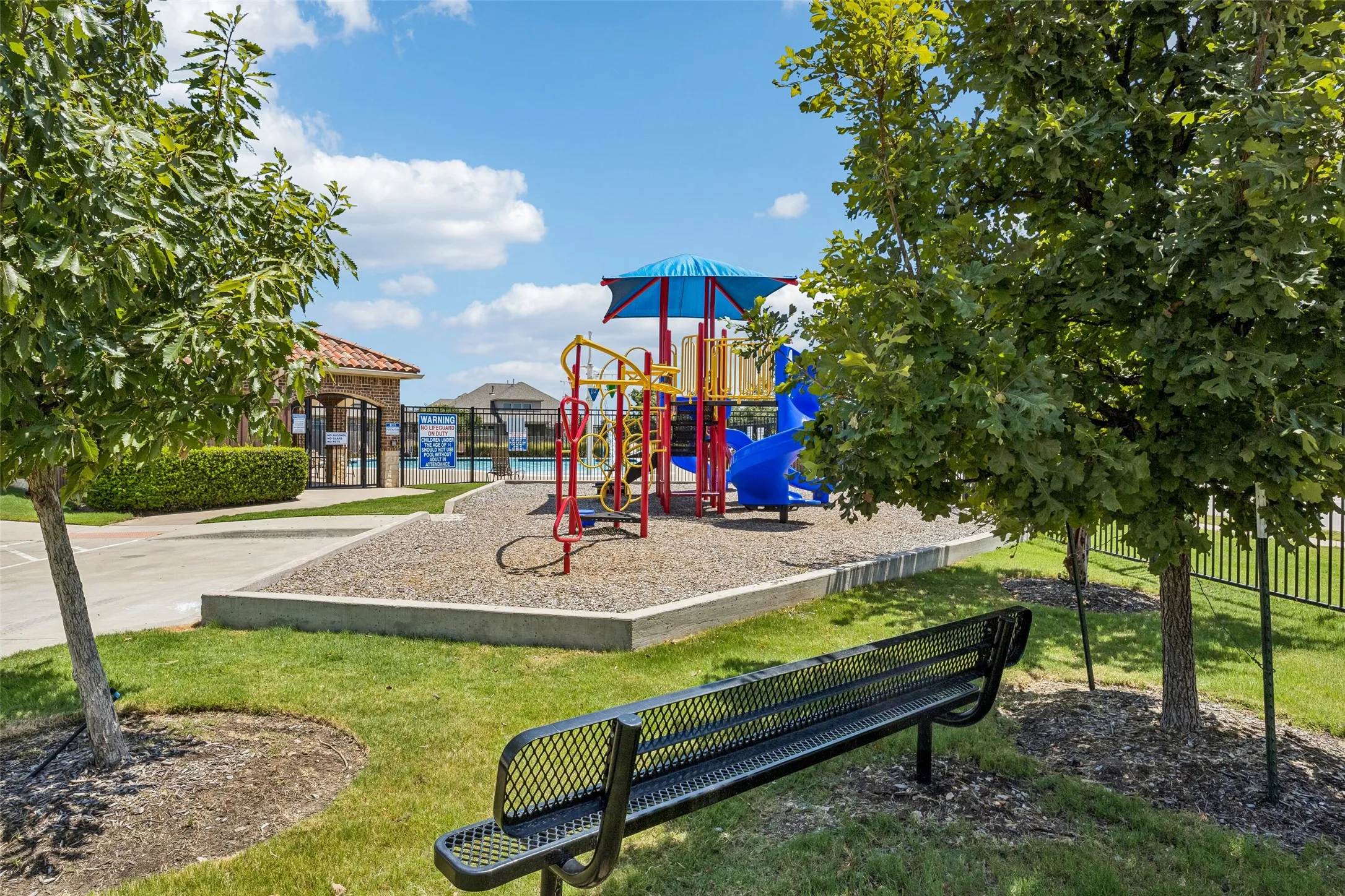 Outdoor playground featuring colorful slides and climbing structures, with a paved walkway, manicured lawn, and mature trees