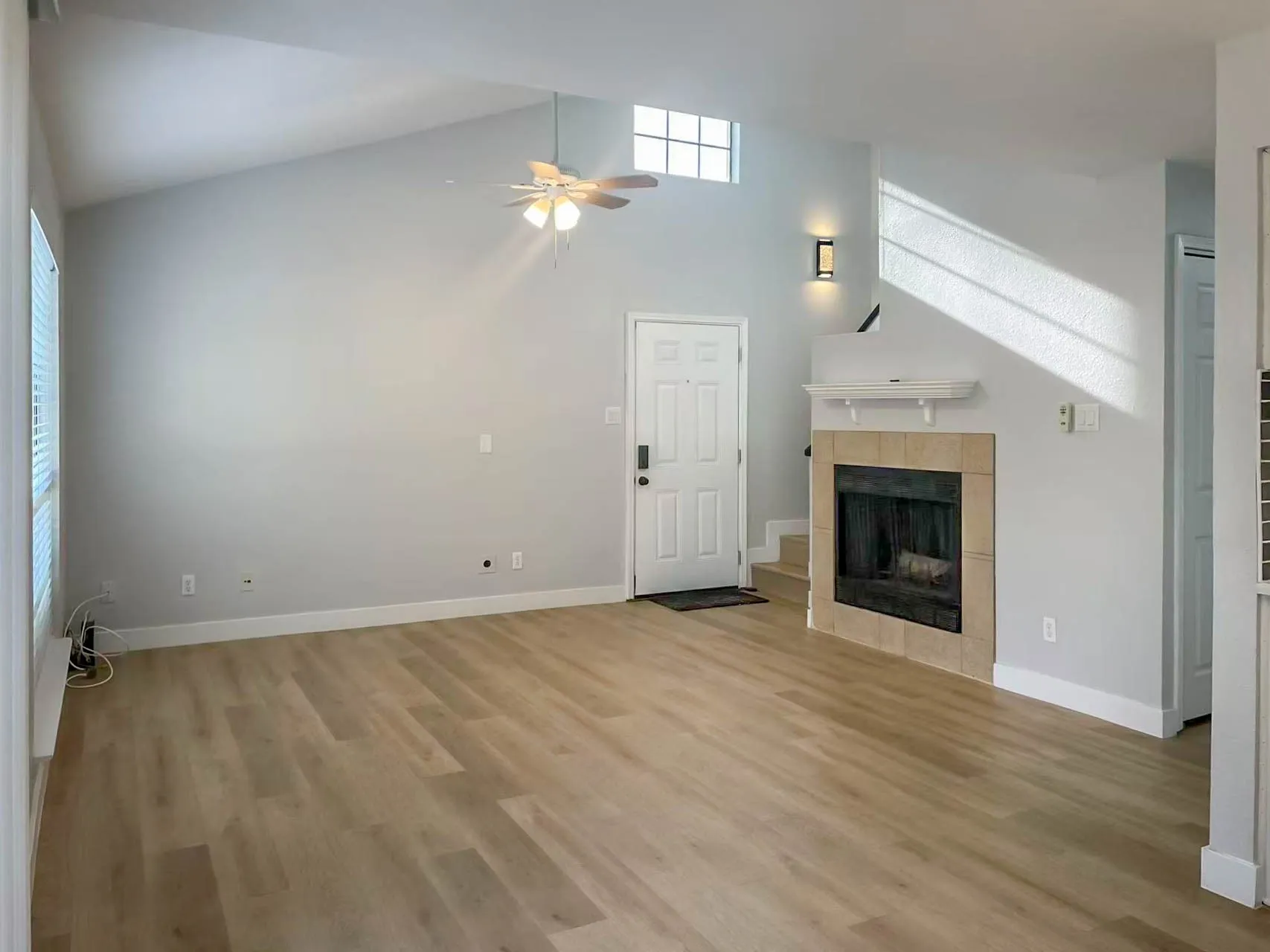 Vaulted ceiling with ceiling fan, wood-finish flooring, tiled fireplace with mantel, transom window, and wall sconce lighting