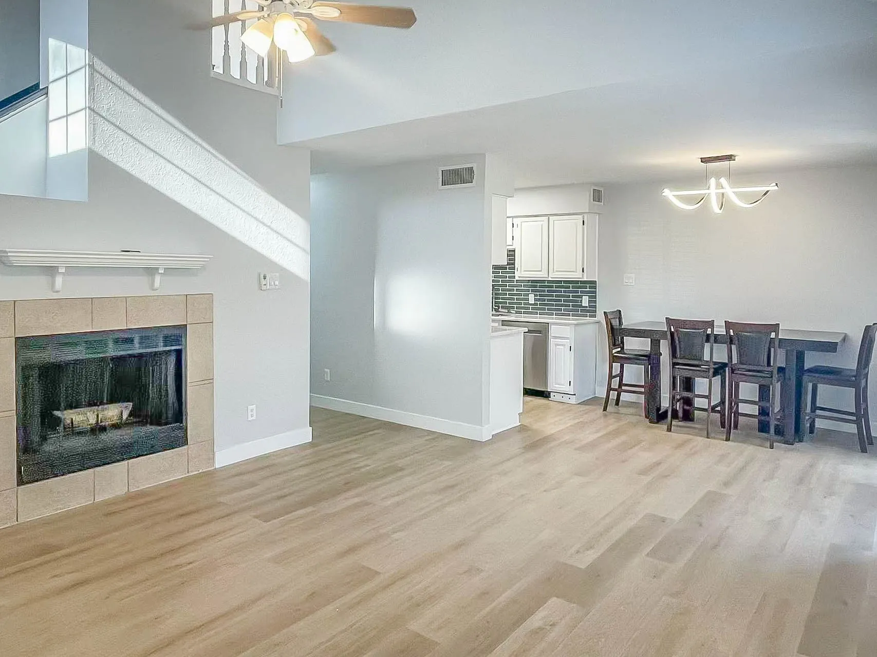 Fireplace with a stone surround and mantel, wood-finish flooring, staircase with white spindles, modern ceiling fan, and kitchen featuring white cabinetry