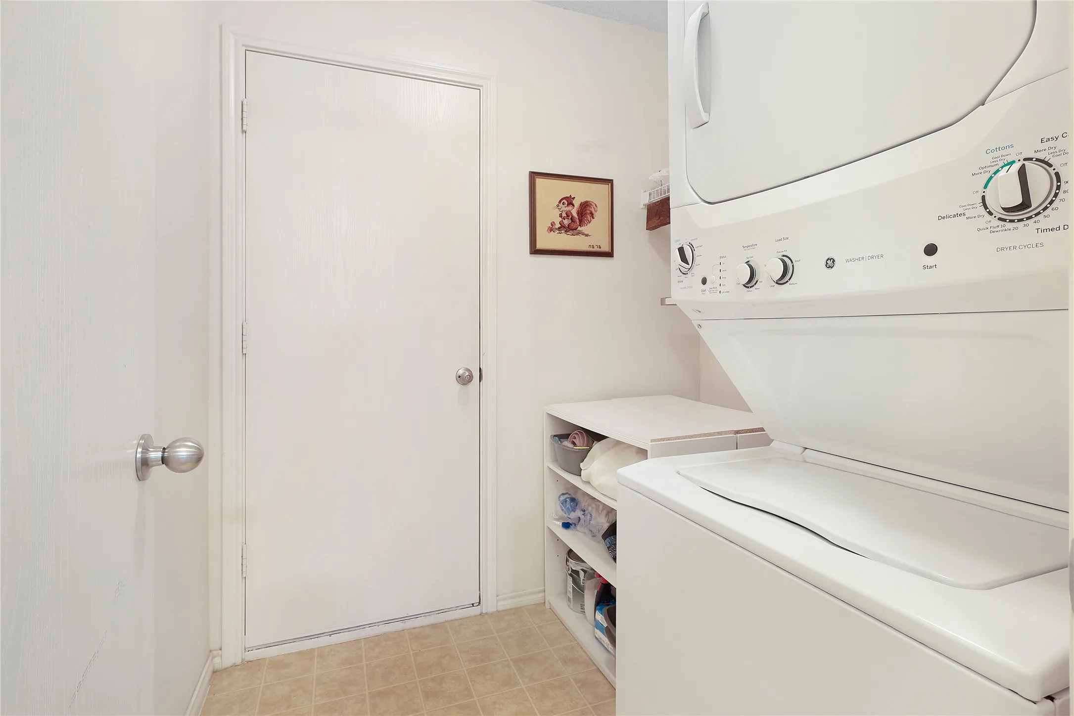 Compact laundry area with a stacked washer and dryer, adjacent built-in shelving, and neutral floor tiling