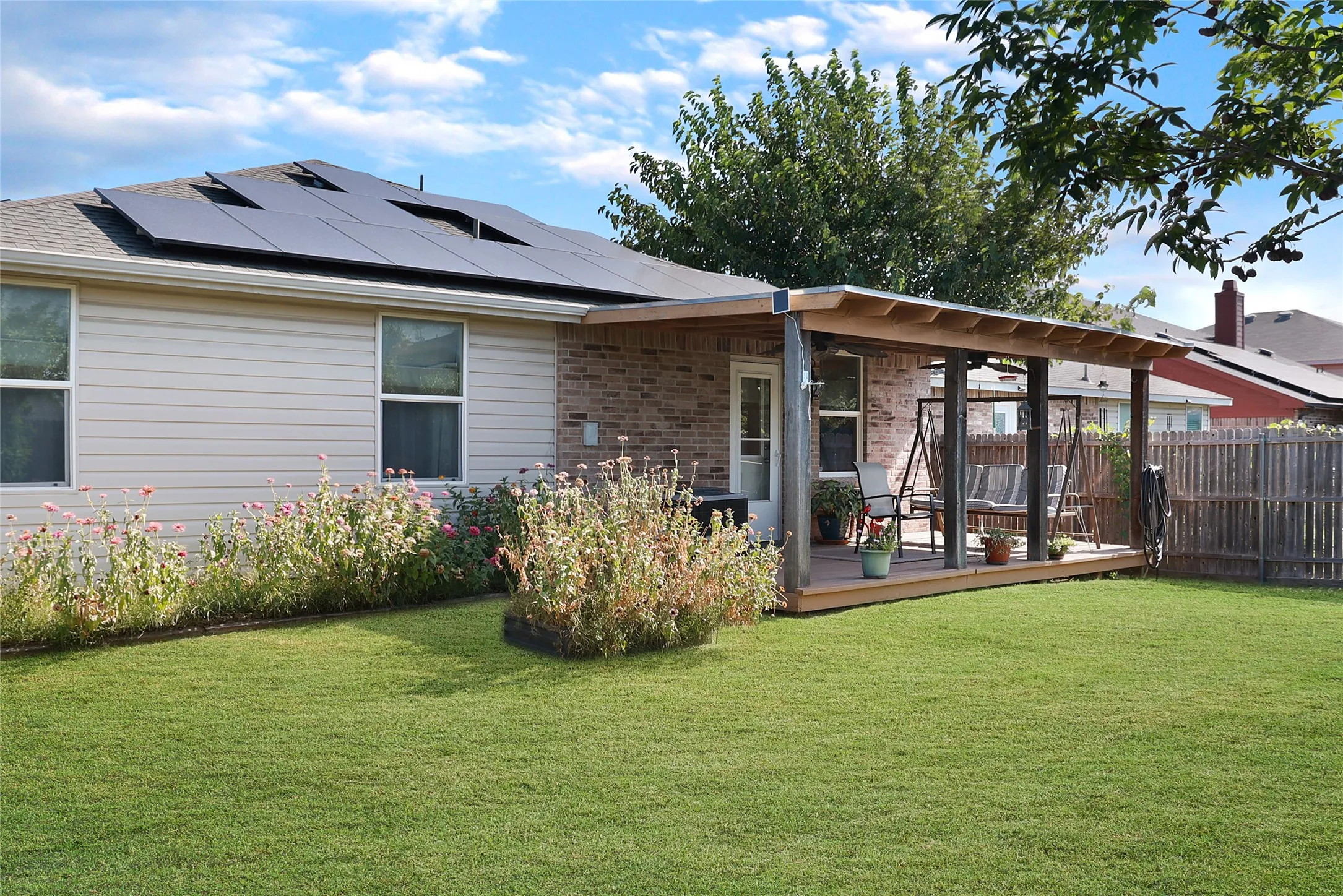 Solar panel array, covered wood-finish deck, brick and siding exterior, fenced backyard, and established landscaping