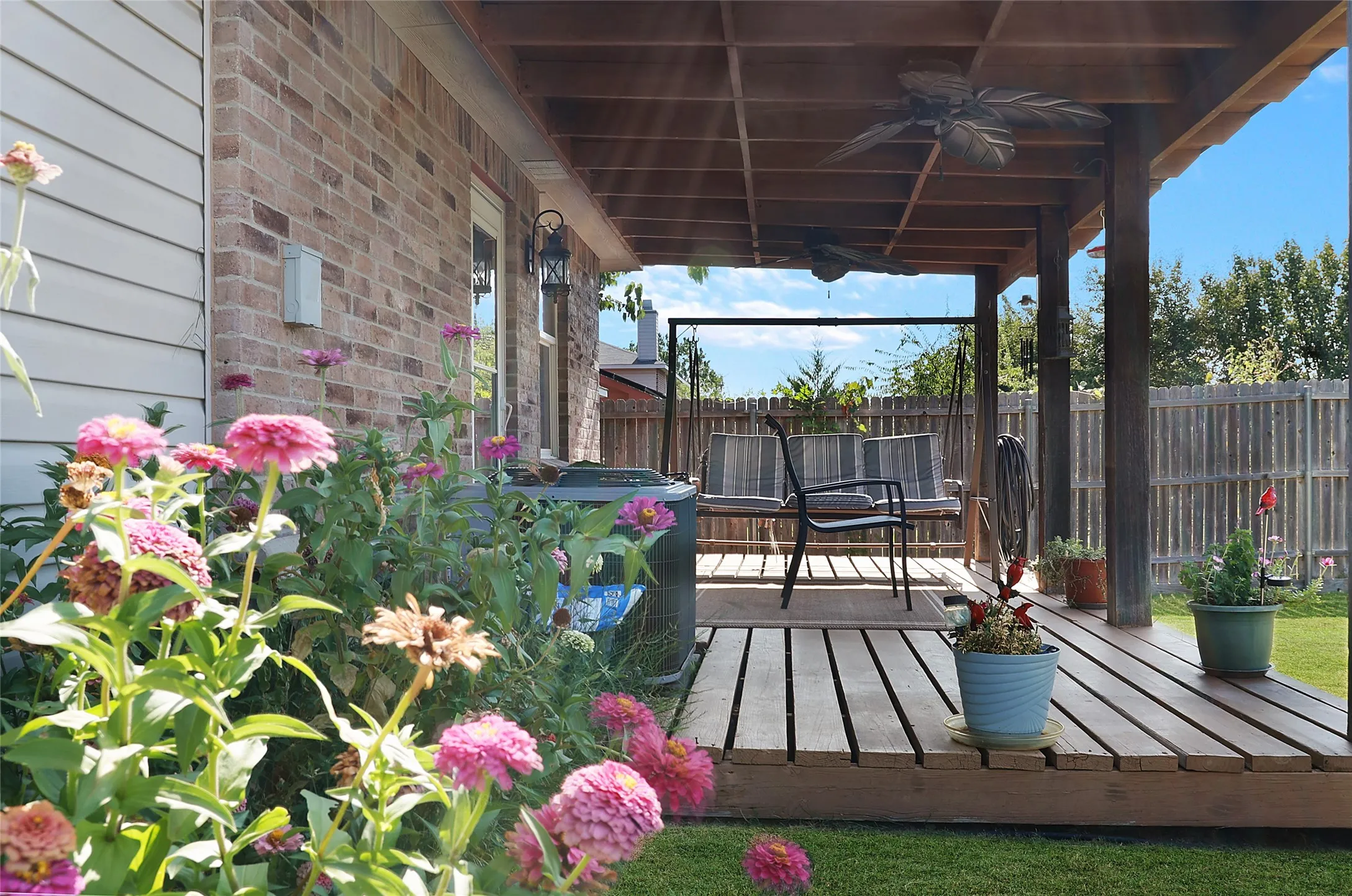Expansive wood-finish deck with a covered patio structure, exterior wall-mounted lighting, and a brick facade