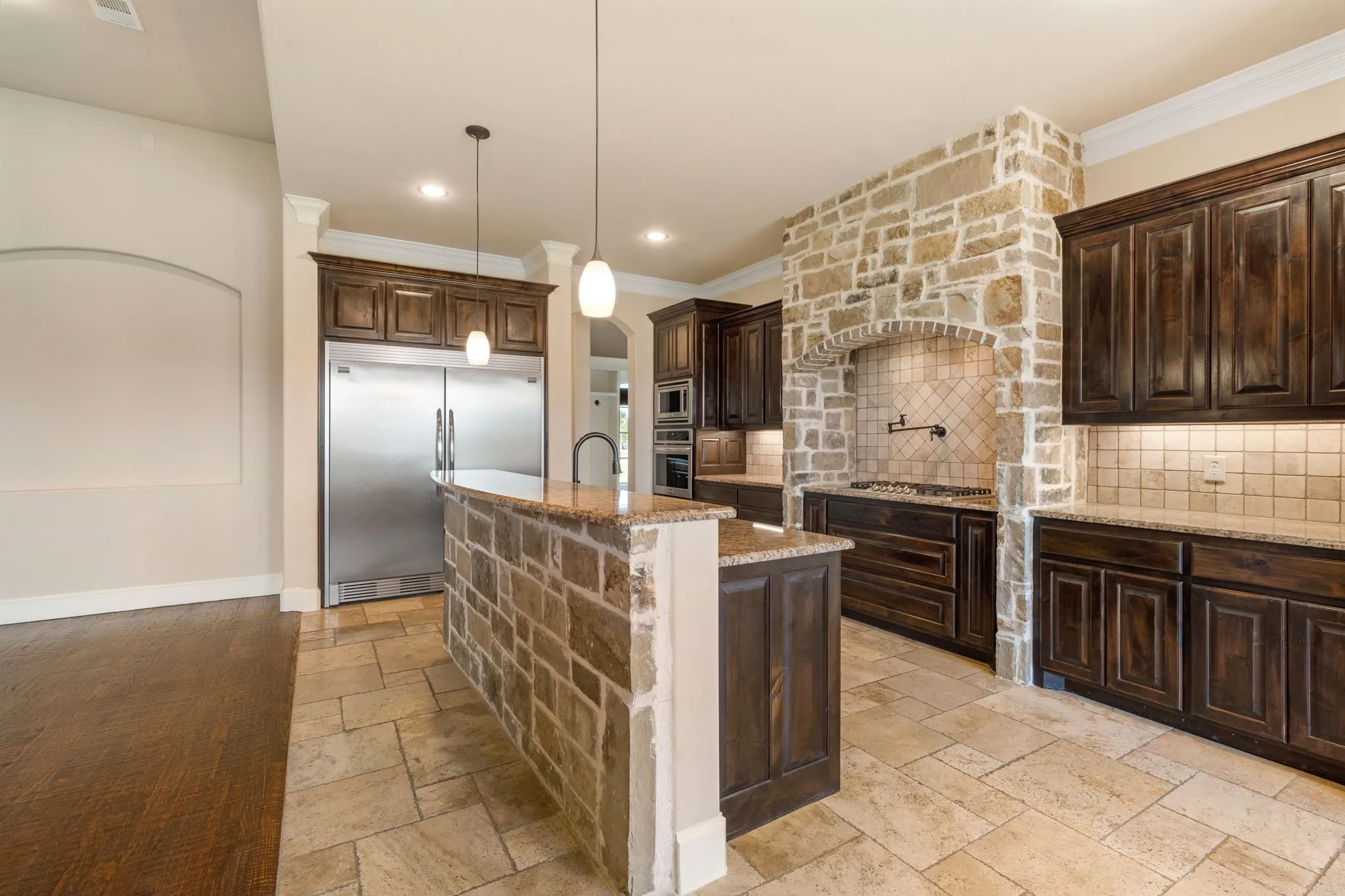 Spacious kitchen featuring dark wood cabinetry, stainless steel refrigerator, brick and stone accents throughout, tile flooring, and a large center island with pendant lighting