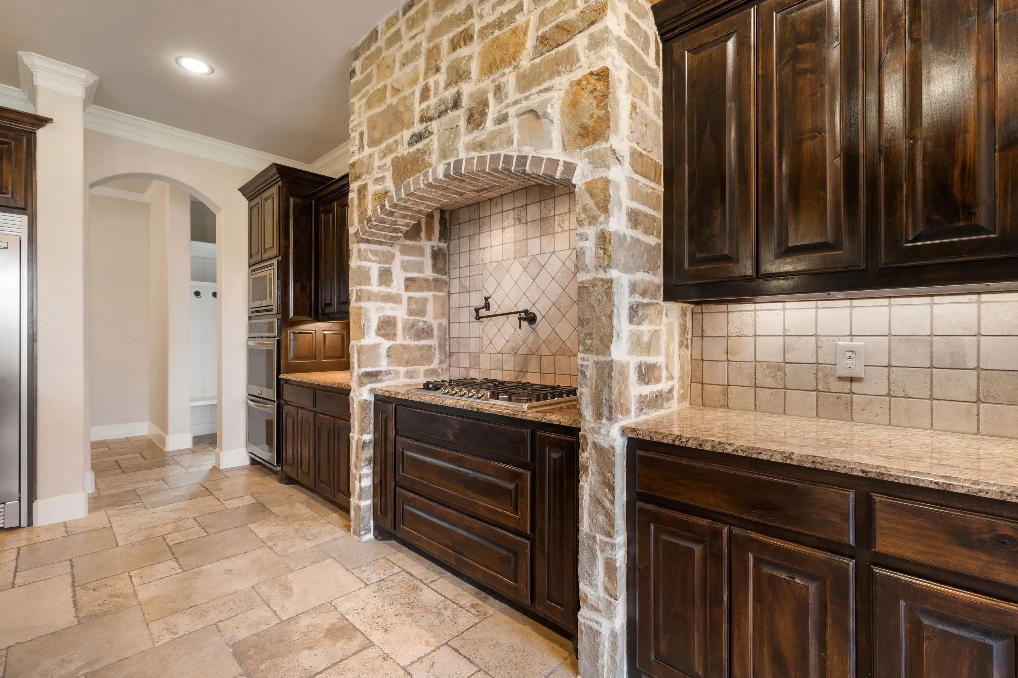 Stone-clad range hood with brick archway, dark wood cabinetry, integrated stainless steel appliances, speckled stone countertops, and tiled flooring