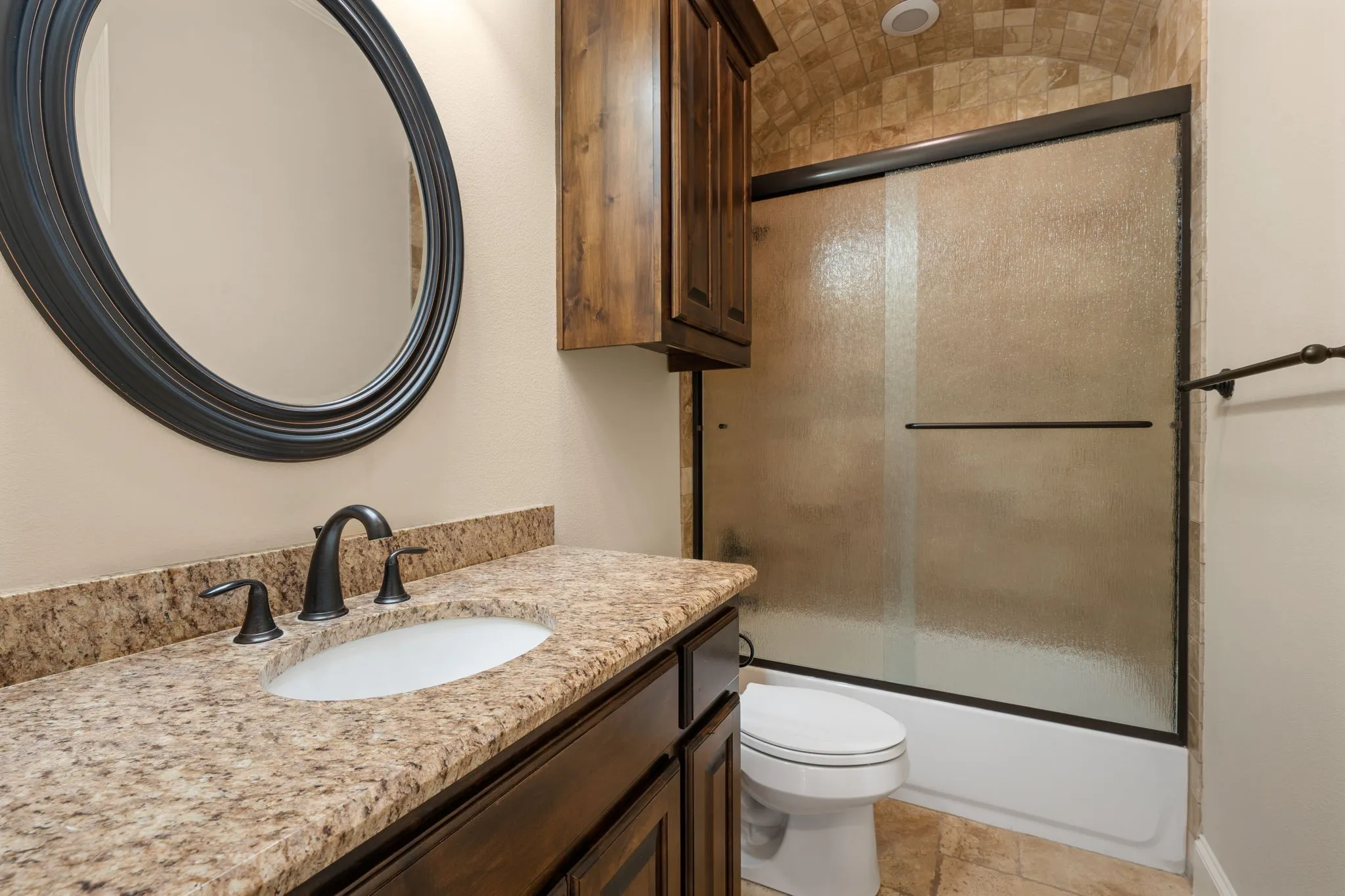 Granite countertop with undermount sink, dark wood vanity cabinetry, oval framed mirror, and a tiled shower/tub combination with sliding frosted glass doors