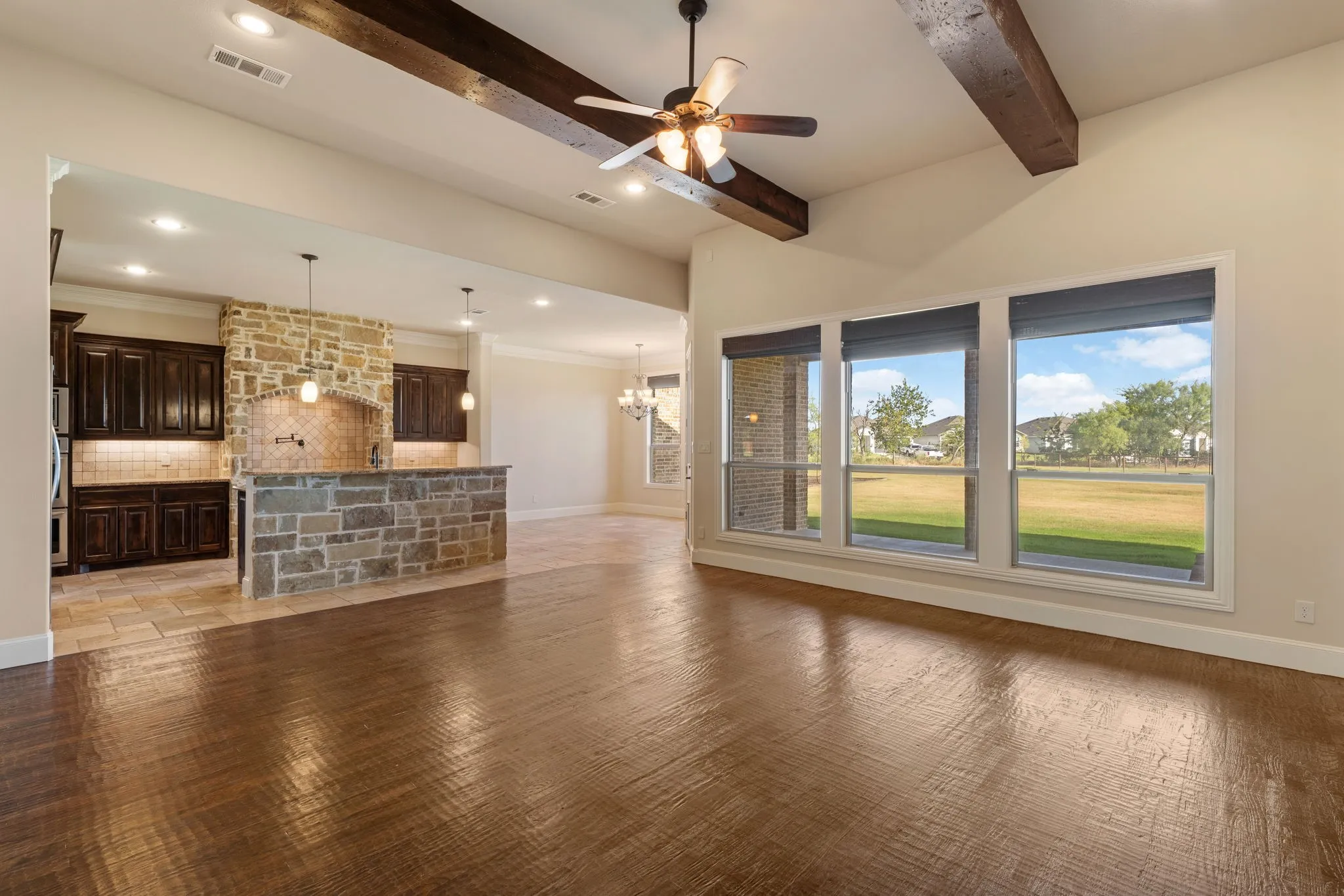 Vaulted ceilings with exposed wood beams, wood-finish flooring, a stone-clad kitchen island, dark wood cabinetry, and large windows with integrated blinds