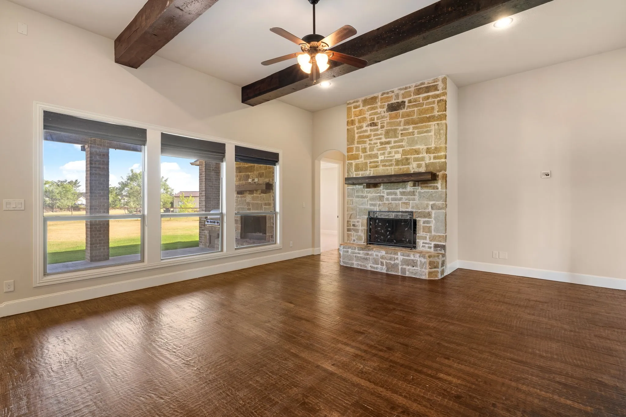 Vaulted ceiling with exposed wood beams, stone-clad fireplace with wood mantel, dark wood-finish flooring, and a ceiling fan