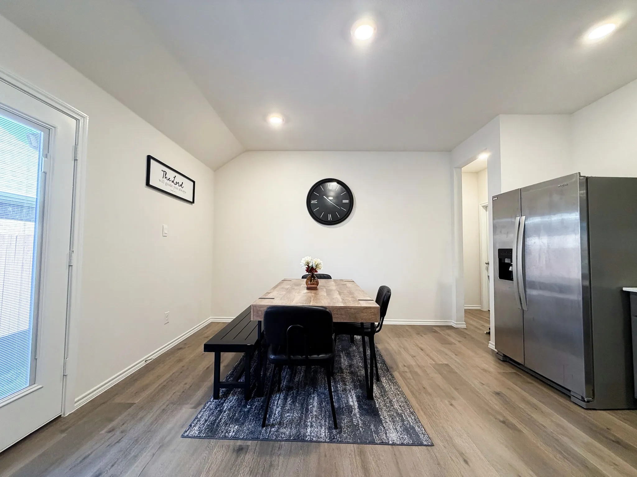 Modern kitchen and dining area featuring stainless steel refrigerator with ice and water dispensers, wood-finish flooring throughout, recessed ceiling lighting, and a spacious dining zone with black metal framing