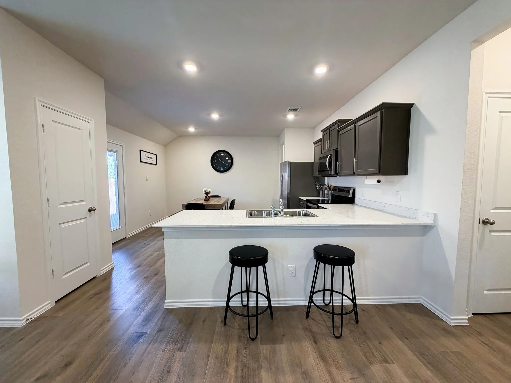 Modern kitchen featuring a white quartz waterfall island with seating for two, black cabinetry with stainless steel appliances, recessed ceiling lighting, and wood-finish flooring throughout the open-concept space