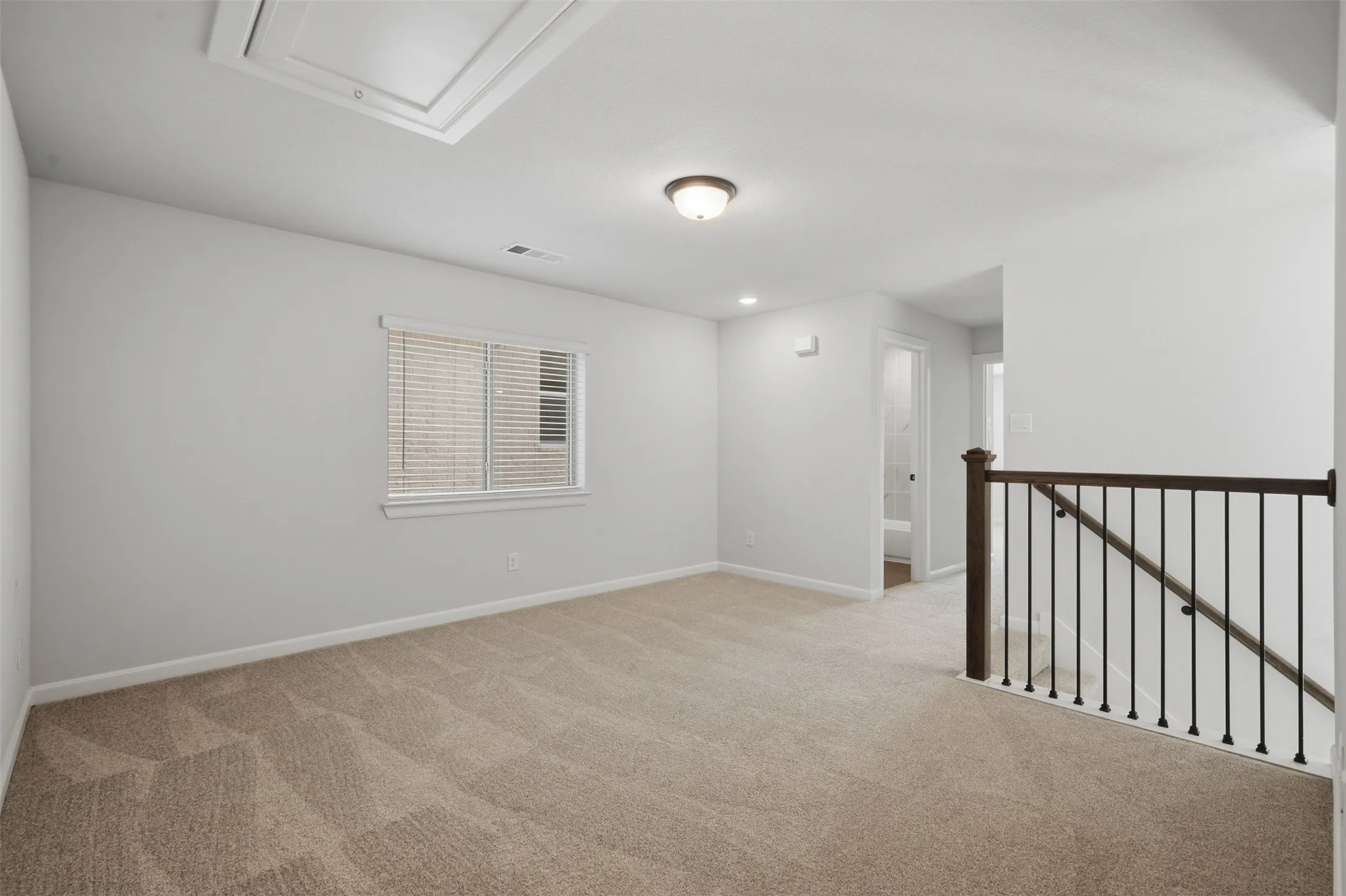 Second-floor landing/loft area featuring plush carpeting, a single window for natural light, a dark wood-and-metal railing, and a modern overhead light fixture, creating a bright and welcoming transition between spaces