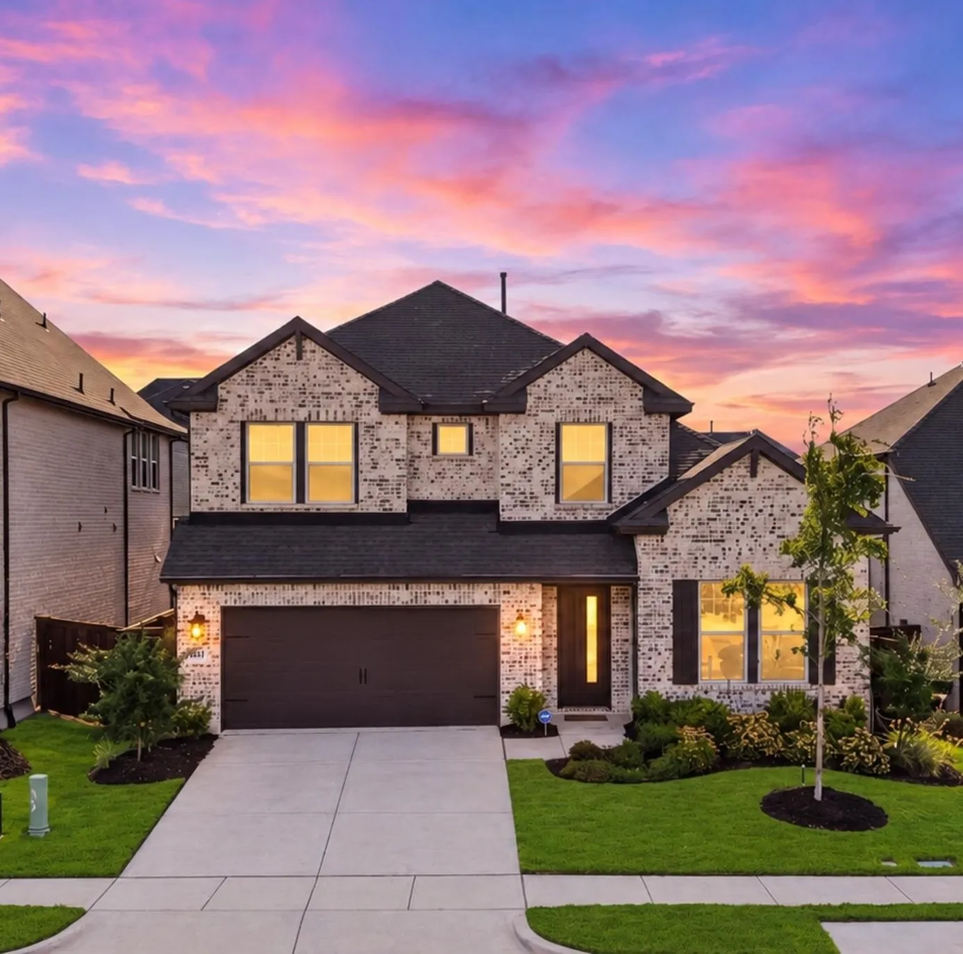 Striking two-story brick and stone residence showcasing a dark architectural roof, illuminated multi-pane windows, and a professionally finished concrete driveway