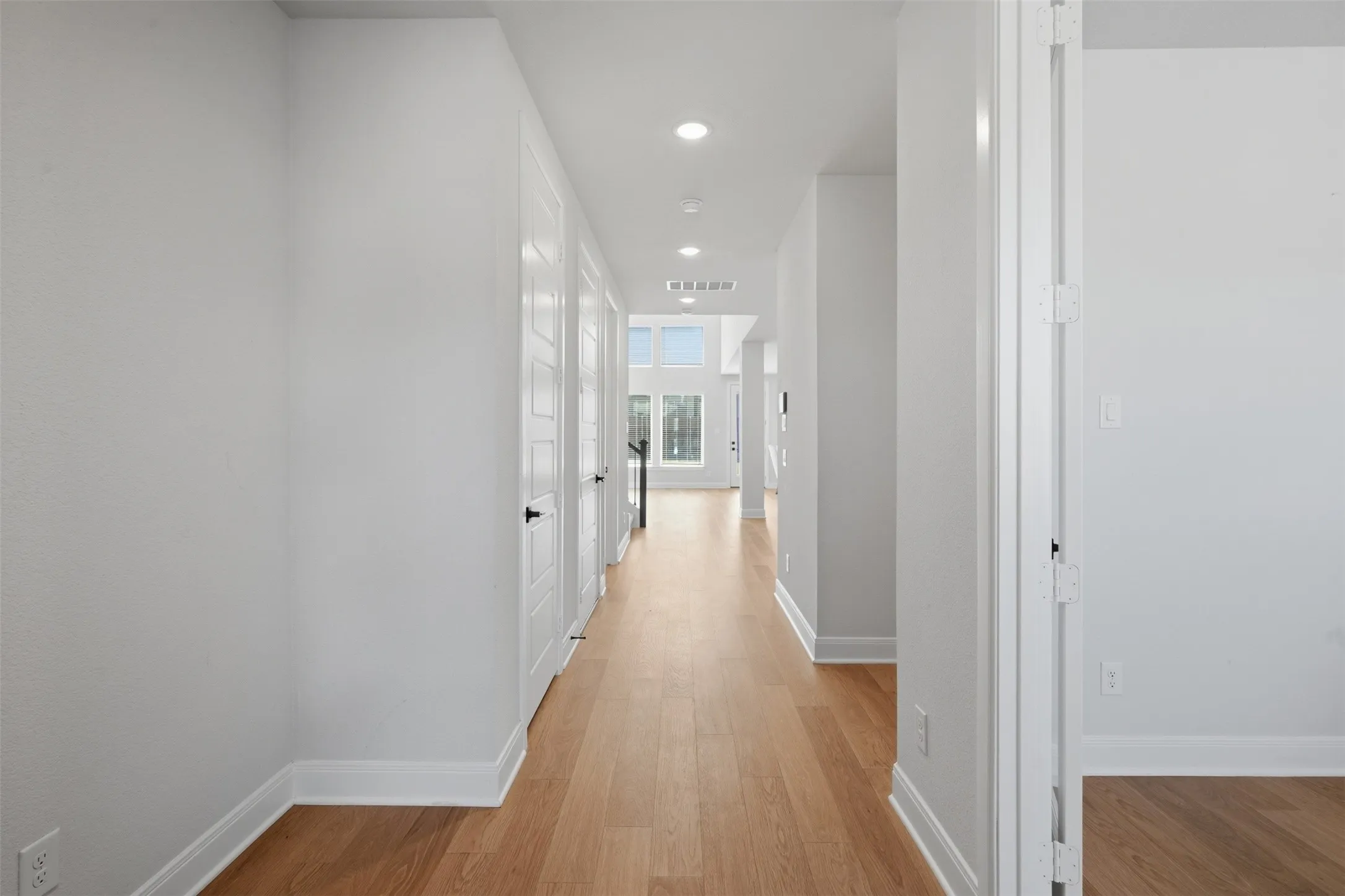 Elegant hallway featuring wood-look flooring, recessed lighting, white doors with matte black hardware, and soaring ceilings accented by transom windows