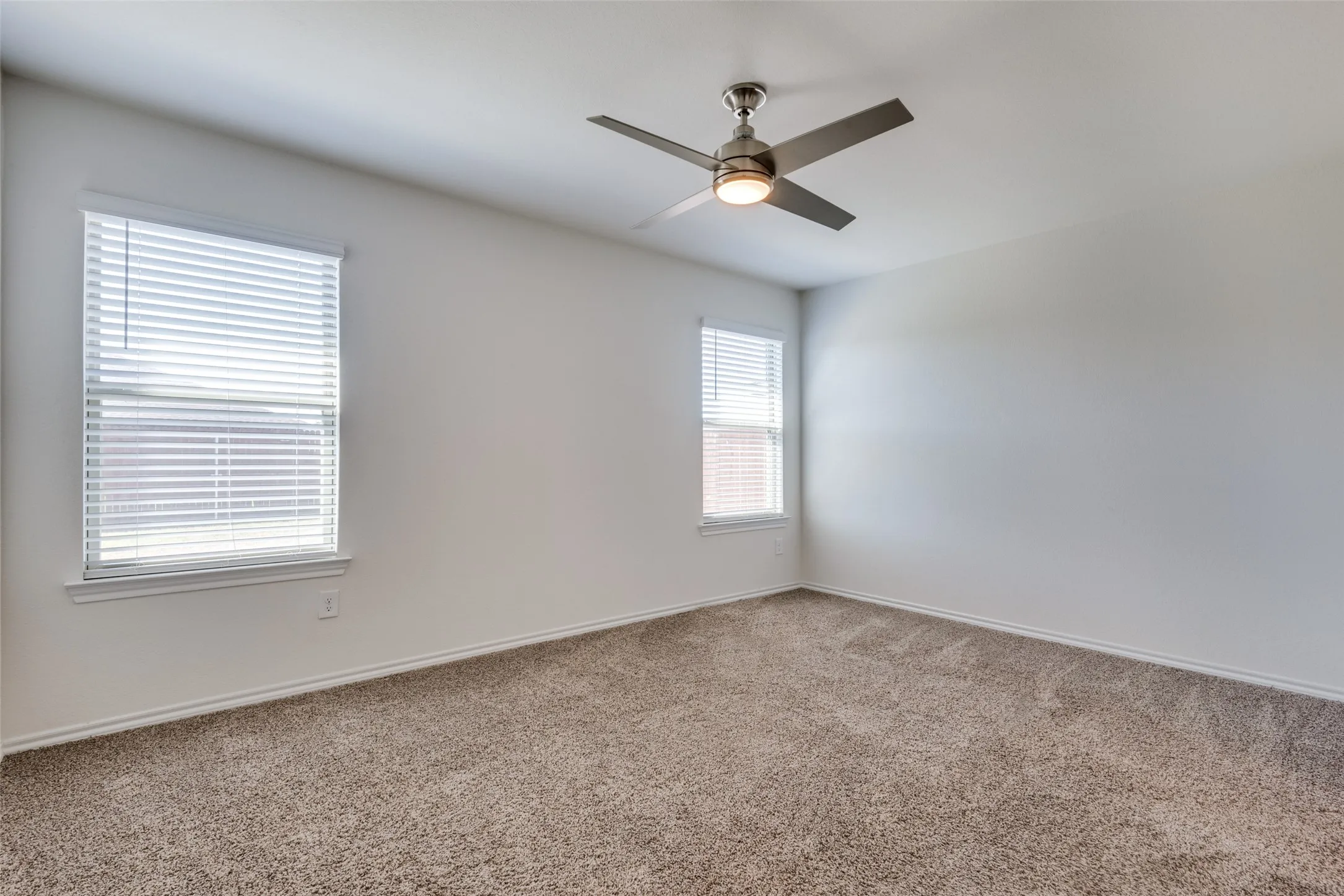 Neutral wall paint, textured carpet flooring, two windows with blinds, and a ceiling fan with integrated lighting
