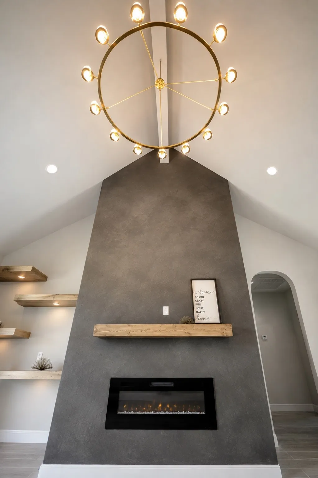 Vaulted ceiling with an overhead light fixture, textured fireplace surround, wood mantel, built-in linear electric fireplace, and built-in wood shelving with integrated lighting