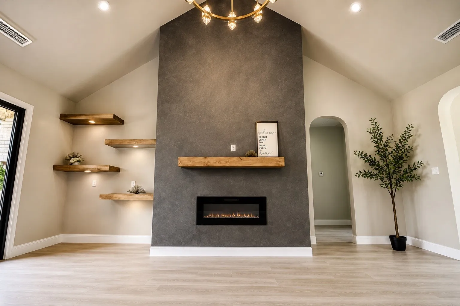 Vaulted ceiling with an ornate chandelier, a floor-to-ceiling accent wall, an electric fireplace with a wood mantle, and wood-finish floating shelves with integrated lighting