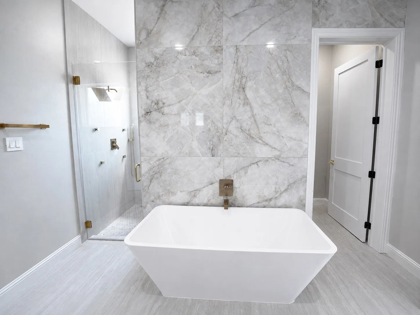 Freestanding soaking tub, floor-to-ceiling marbled tile, glass-enclosed shower, wood-finish flooring, and a modern two-panel door
