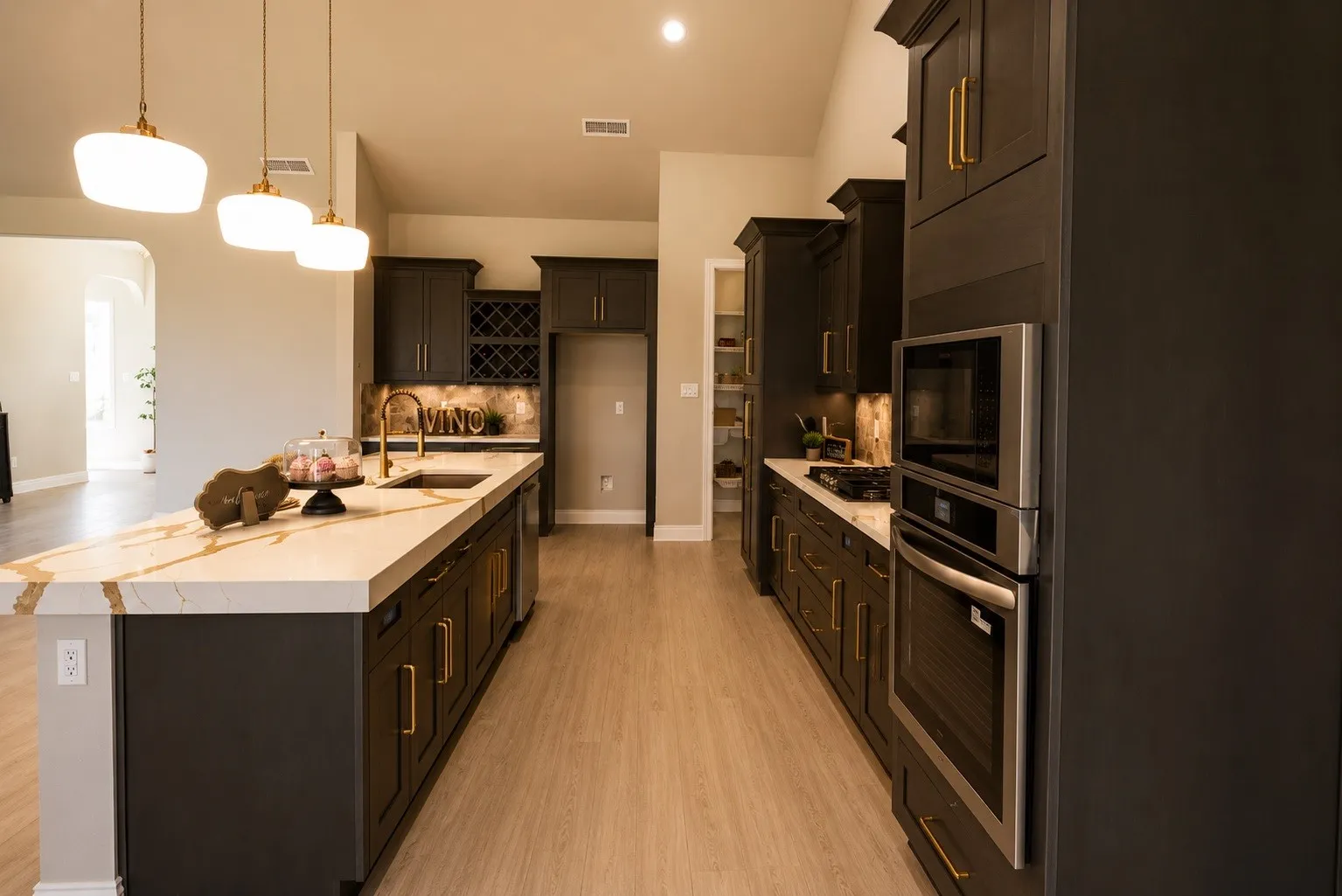 Spacious kitchen featuring an extended island with a light-toned countertop, dark cabinetry with gold-toned hardware, stainless steel built-in appliances, and light wood-finish flooring