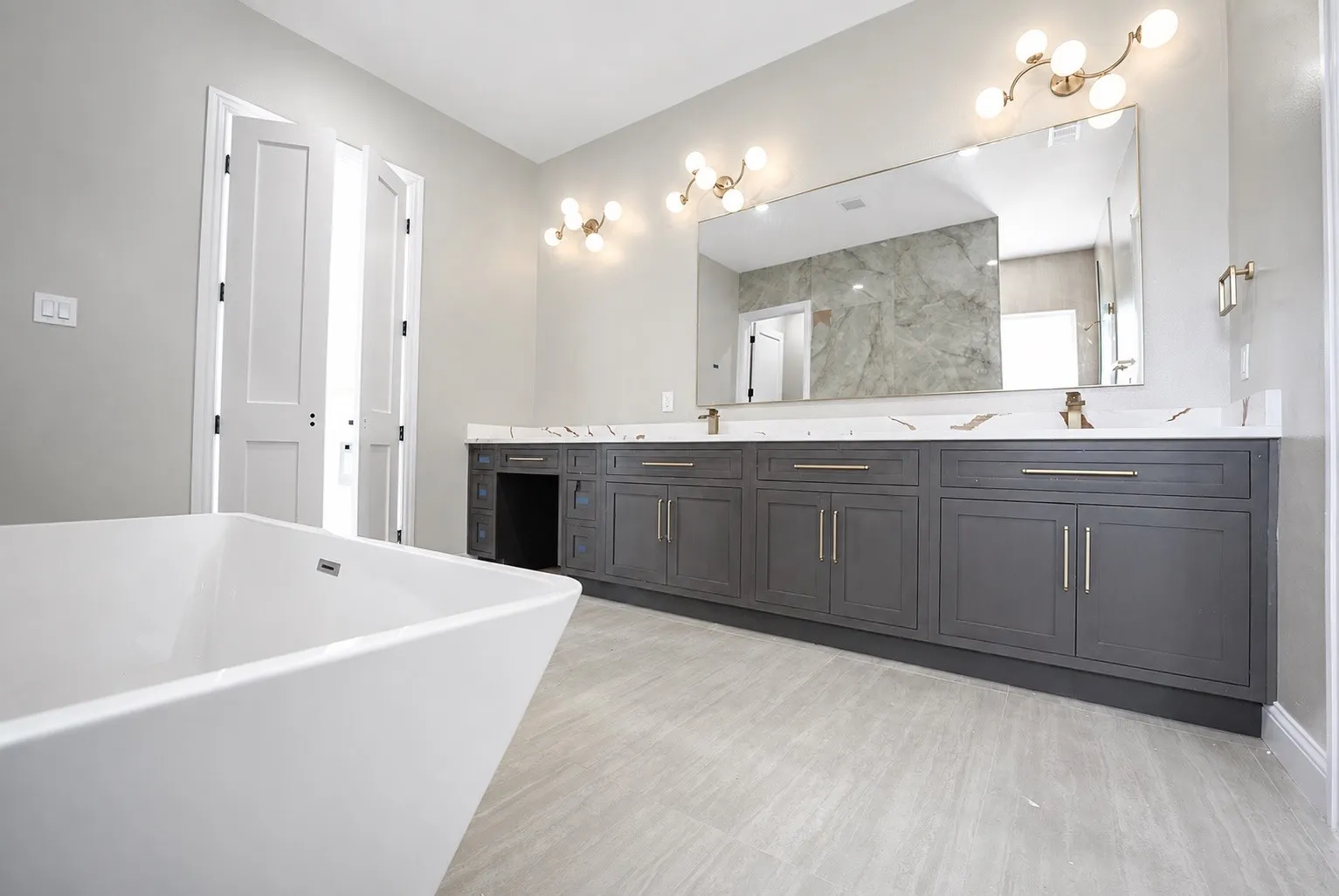 Freestanding soaking tub, double vanity with gray cabinetry, white countertops, large mirror, and tiled flooring