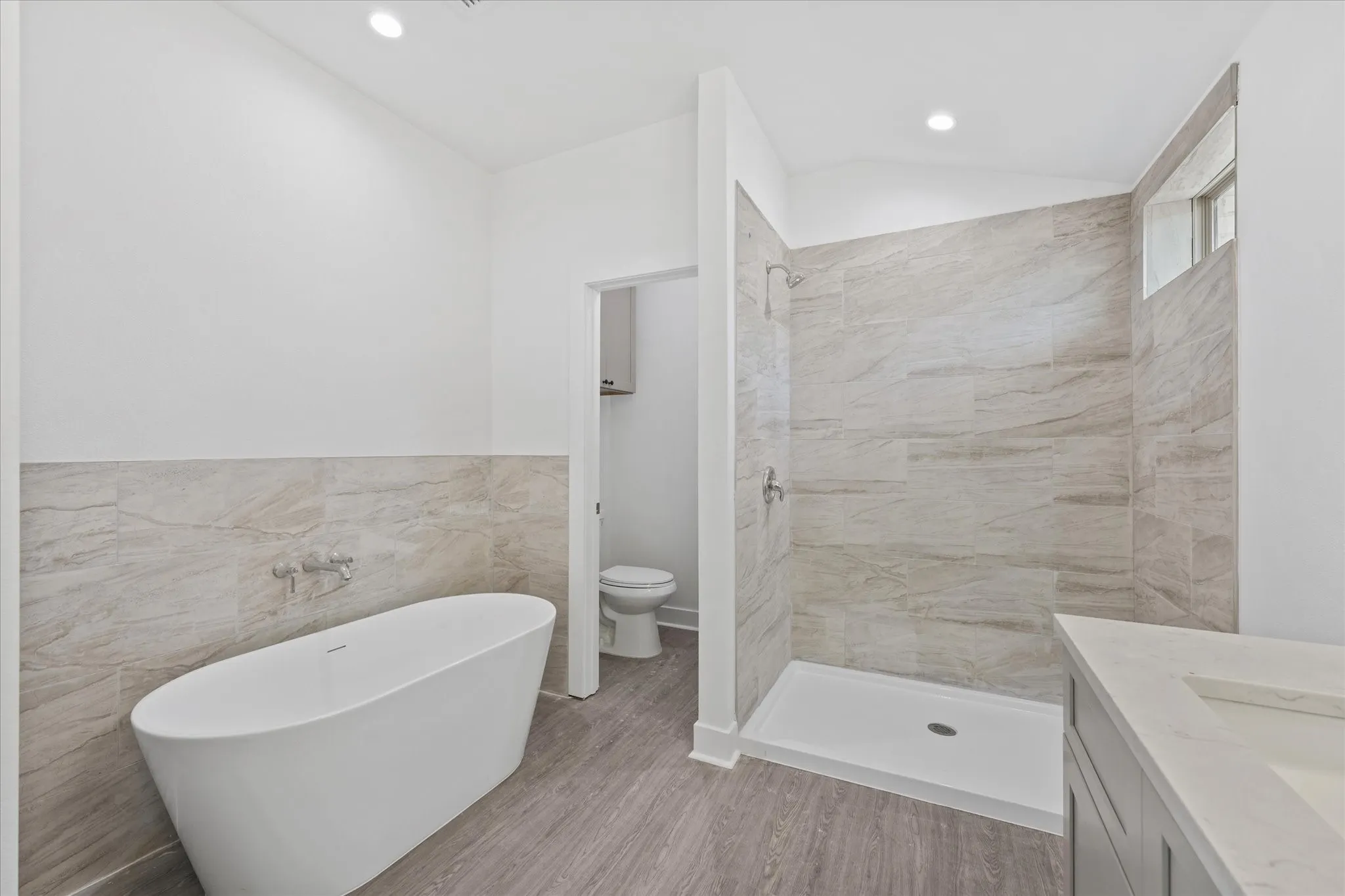Freestanding soaking tub, walk-in shower with tiled walls, built-in vanity with integrated sink, and wood-finish flooring