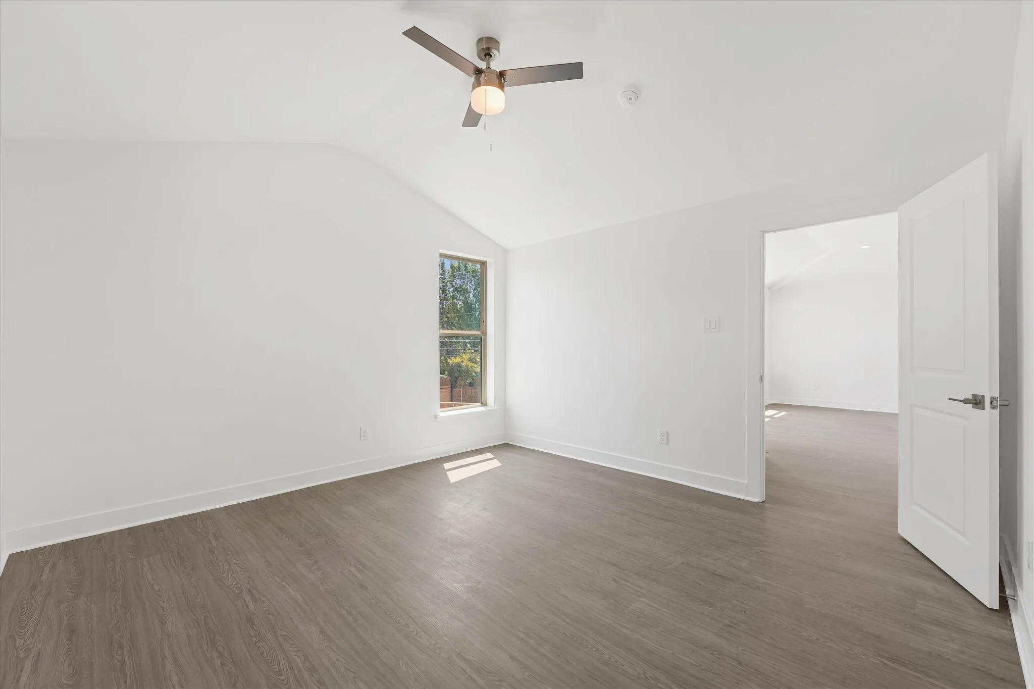Vaulted ceiling, wood-finish flooring, ceiling fan with integrated lighting, white interior door with brushed nickel hardware, and a double-hung window