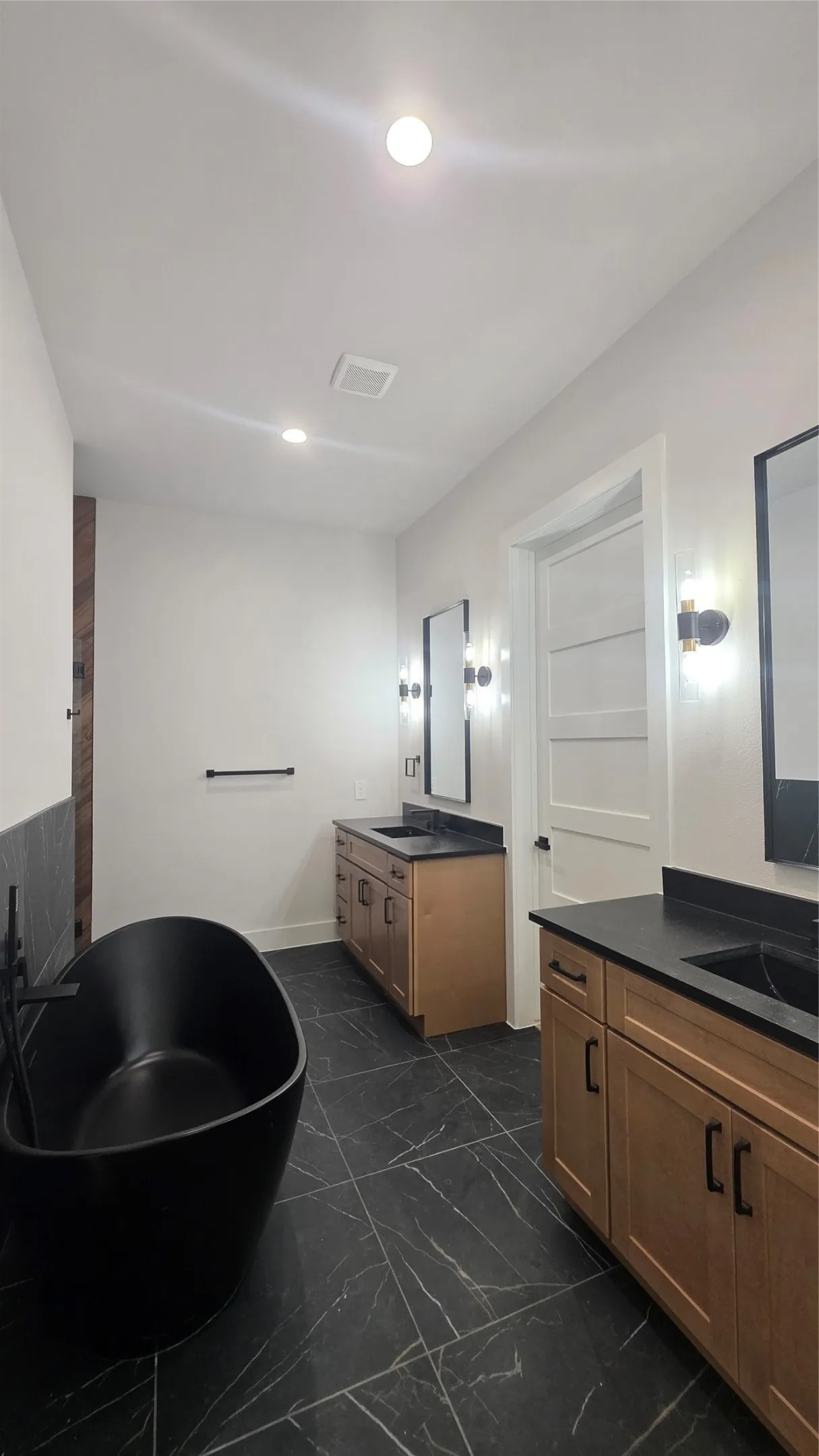 Freestanding black soaking tub, dual wood-finish vanities with black countertops, and black-veined tile flooring