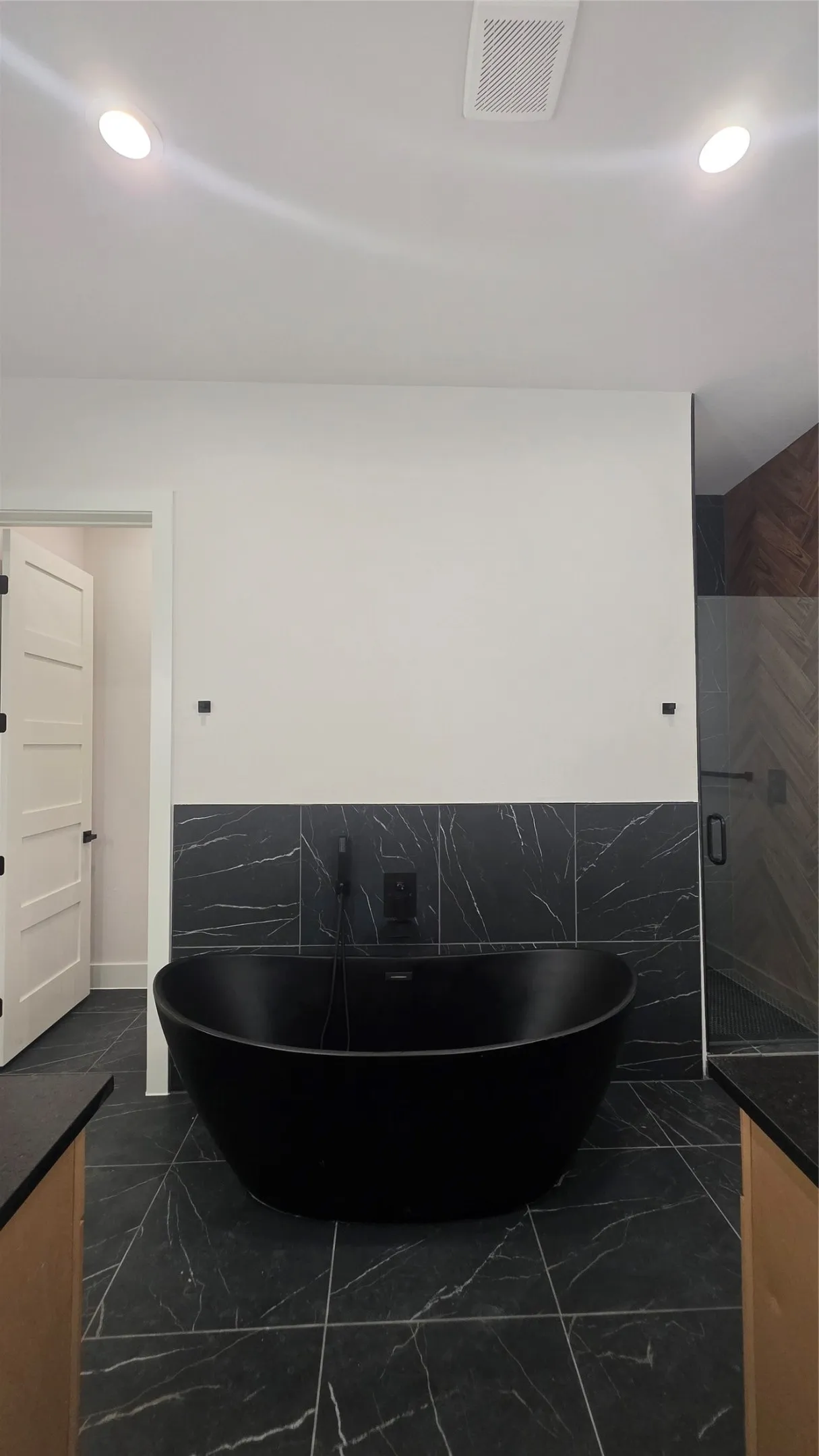Matte black freestanding bathtub, dark marbled tile flooring, dark marbled accent wall, black wall-mounted faucet, and recessed lighting