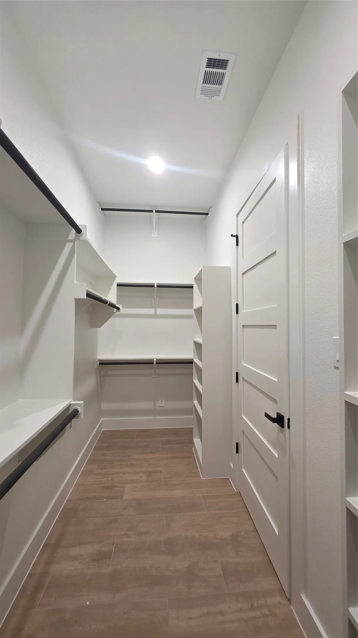 Walk-in closet featuring wood-finish flooring, built-in shelving, black hanging rods, and a white interior door with black hardware