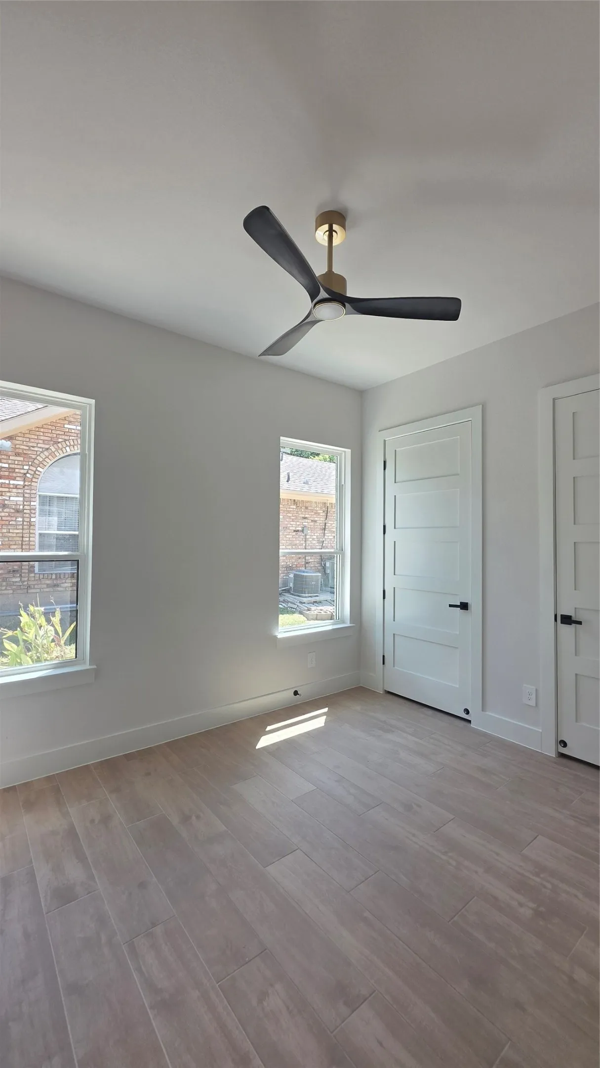 Light wood-finish flooring, white walls, two windows, and a ceiling fan with dark blades