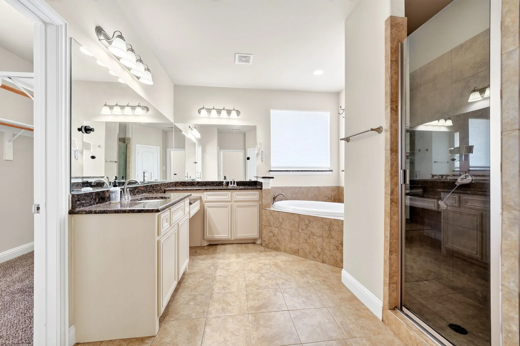 Dual vanity with dark stone countertops, built-in soaking tub, separate glass-enclosed shower, and tiled flooring