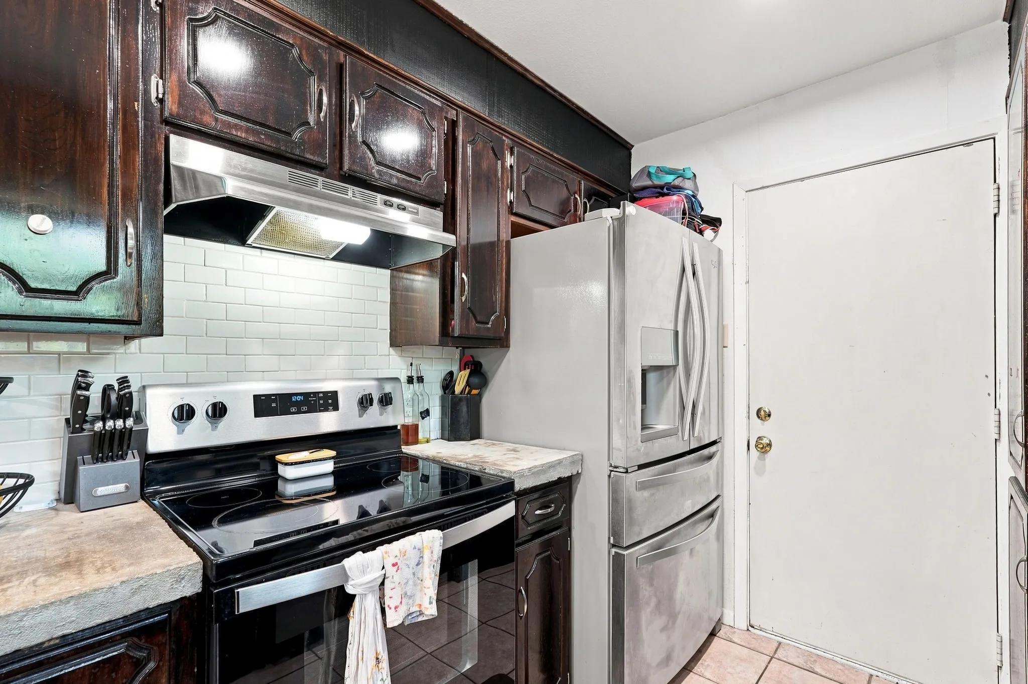 Dark wood-finish cabinetry, stainless steel range hood, white subway tile backsplash, stainless steel electric range, and light-toned countertops