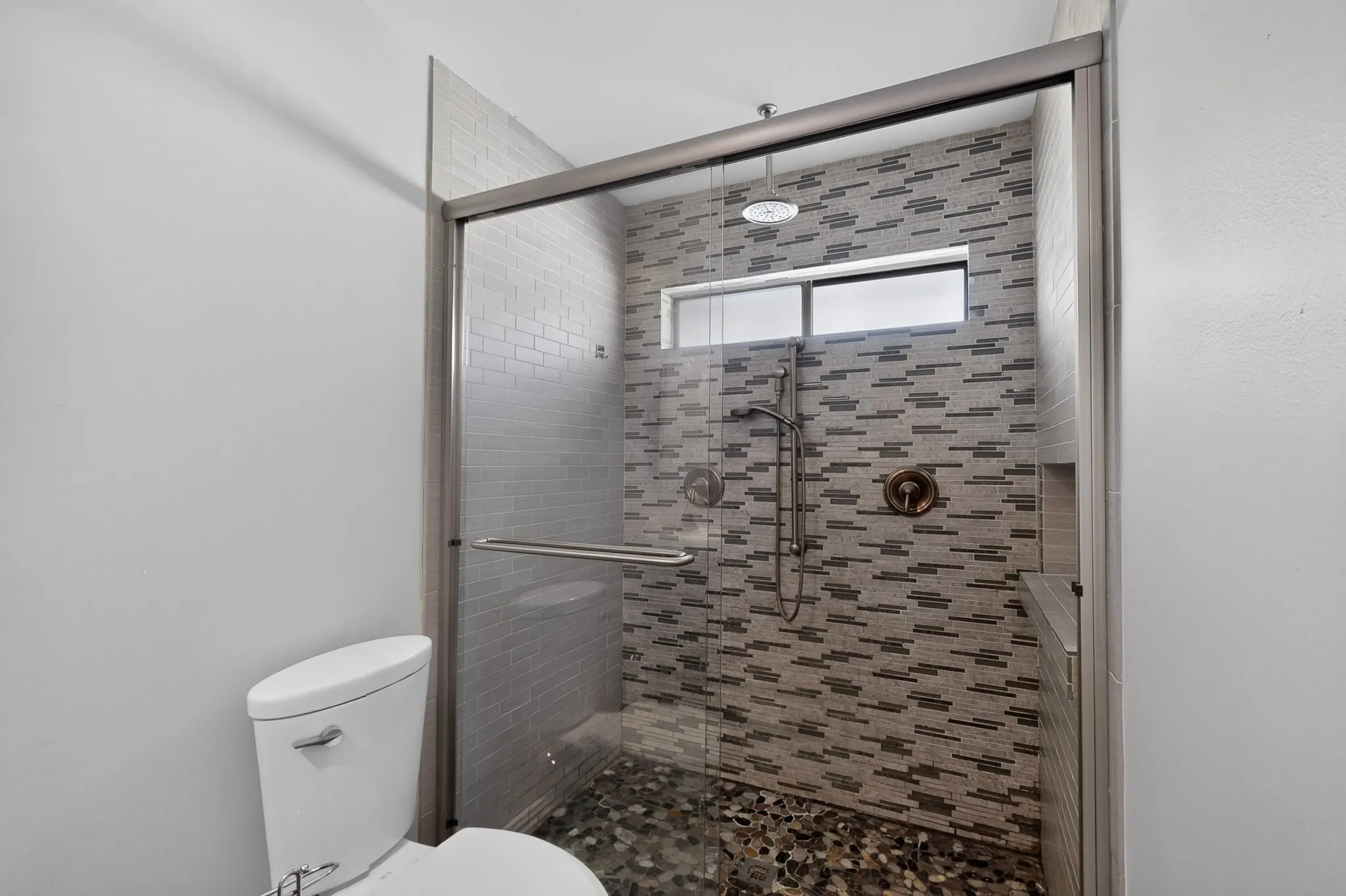Glass-enclosed shower with a rain shower head, handheld shower, stone pebble flooring, and a tiled accent wall