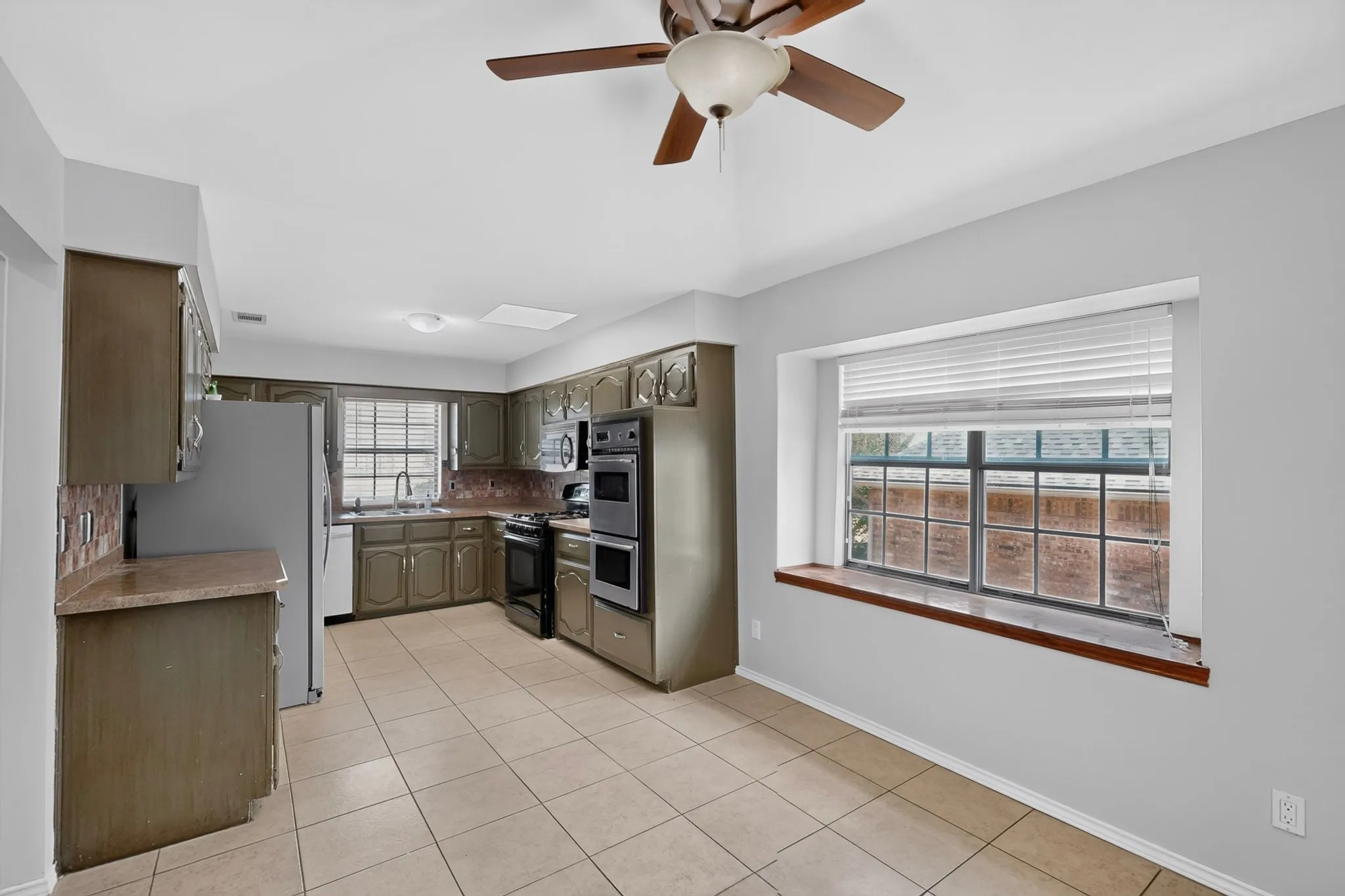 Spacious kitchen featuring stainless steel appliances, wood-finish cabinetry, tile flooring, and abundant natural light from large windows with wood-trimmed sills and window seat