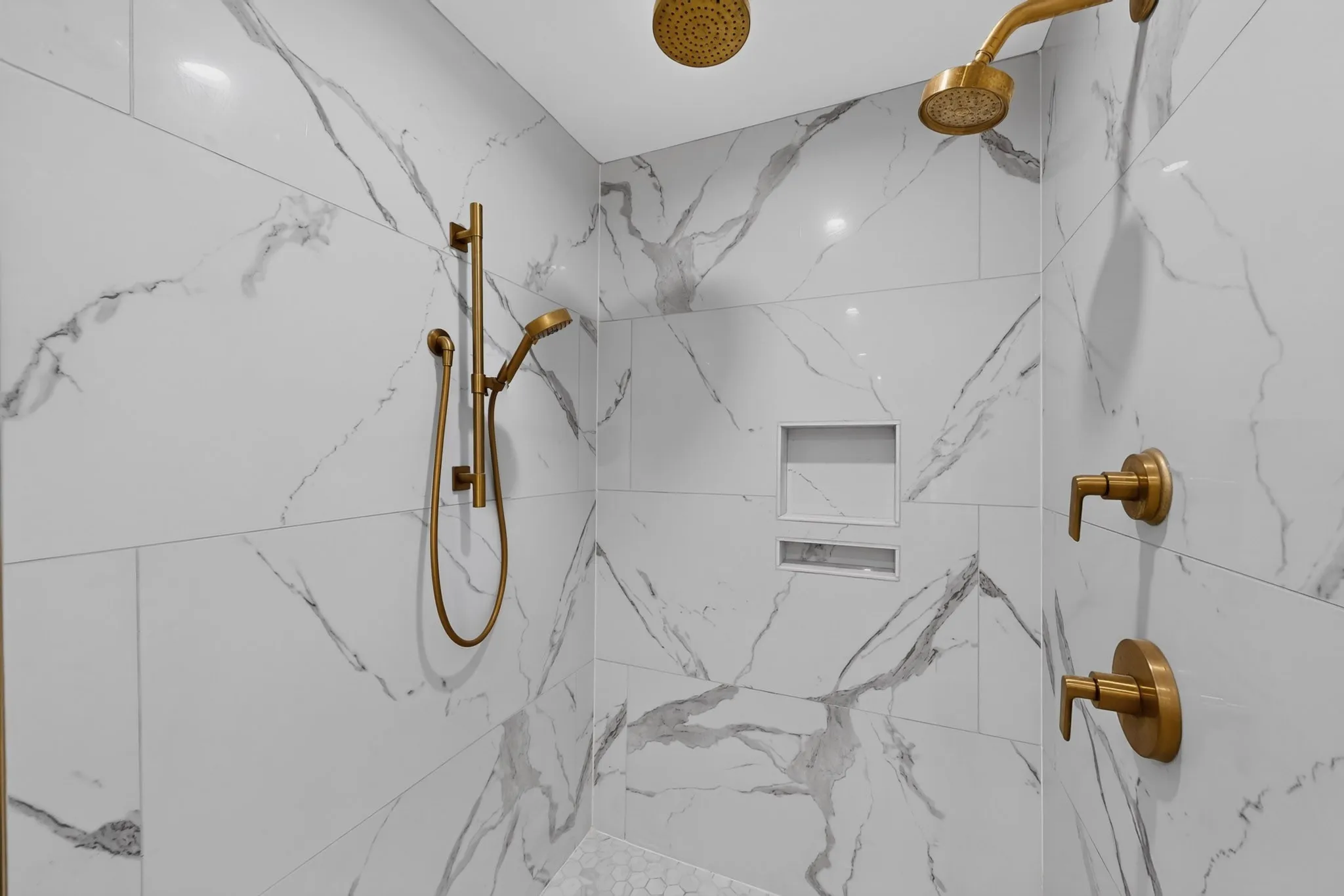 White and grey veined tile shower walls, dual shower heads, built-in recessed shelving, gold-finish fixtures, and a handheld shower