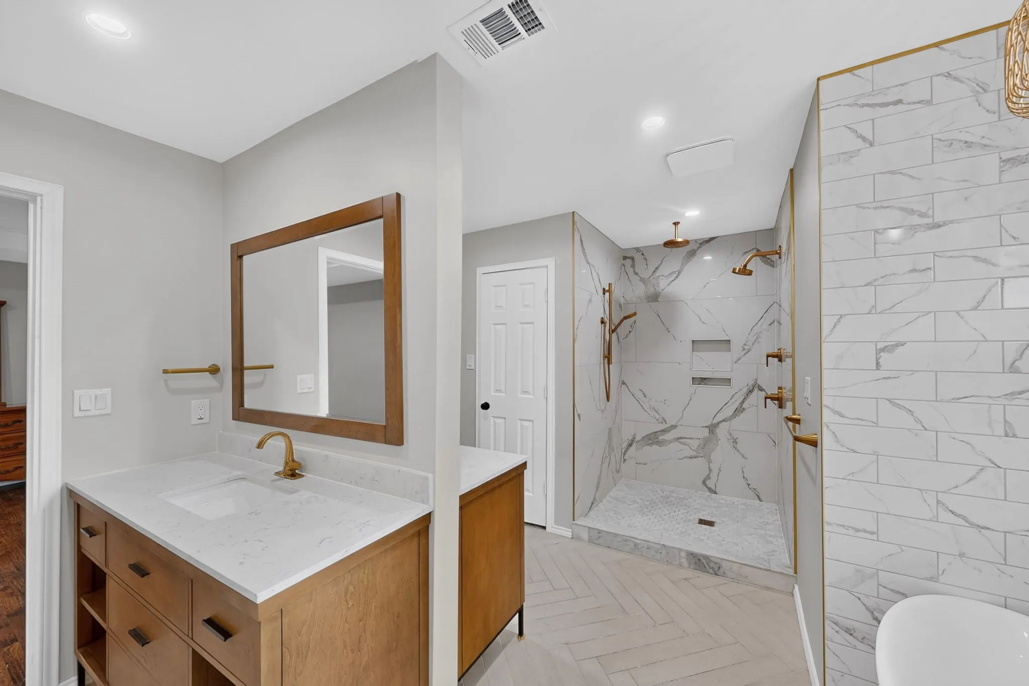 Dual shower system, wood-finish double sided vanity with countertop, framed mirror, herringbone pattern flooring, and white tiled shower walls