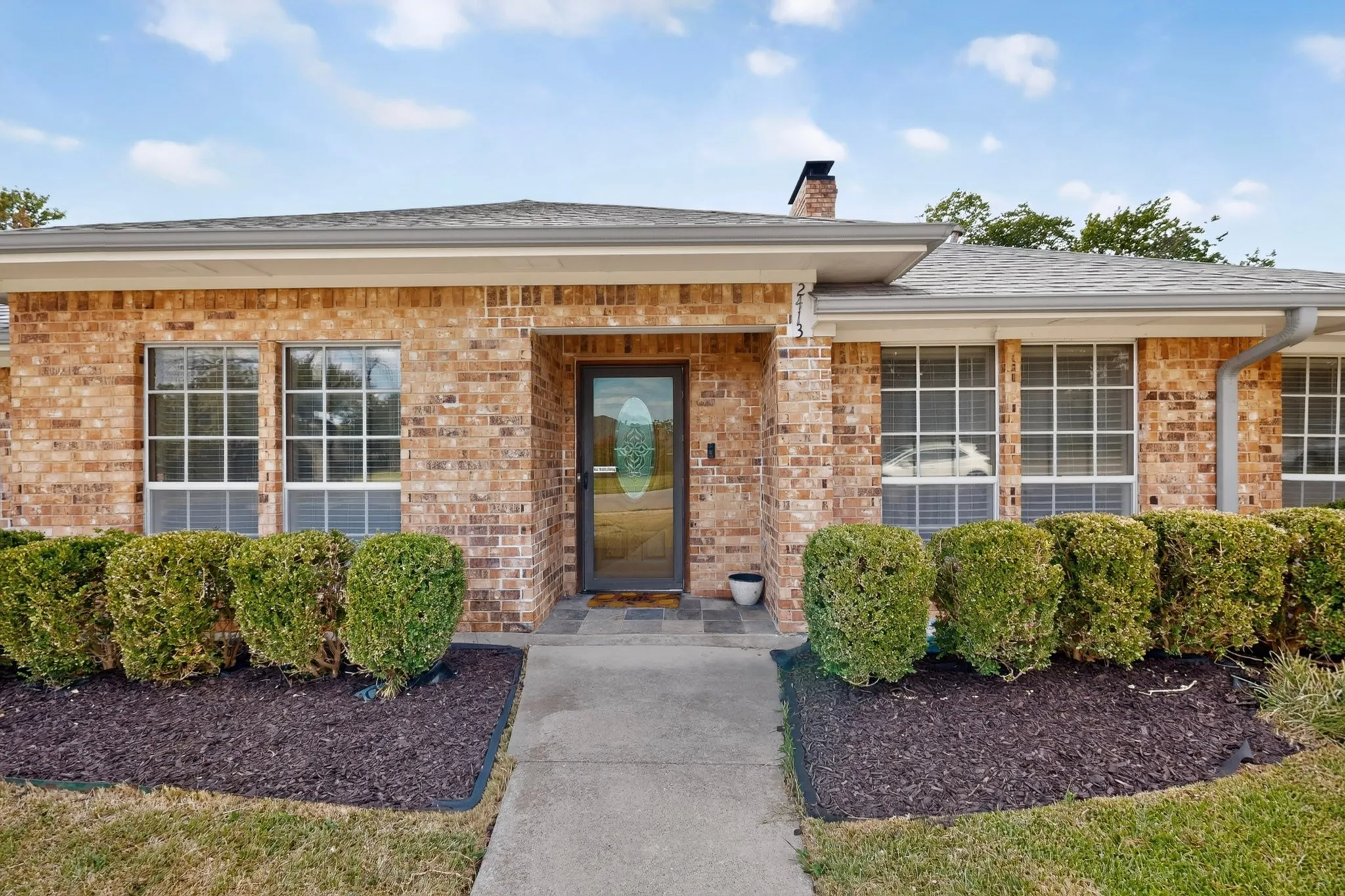 Brick exterior with multiple windows, a central entry door with decorative glass, and a concrete walkway