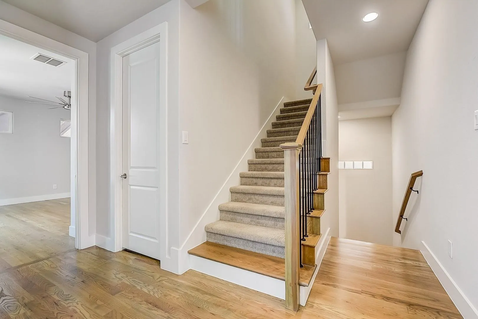 Hardwood-finish flooring, carpeted stairs, recessed lighting, white trim, and a staircase with a wood handrail and black iron balusters