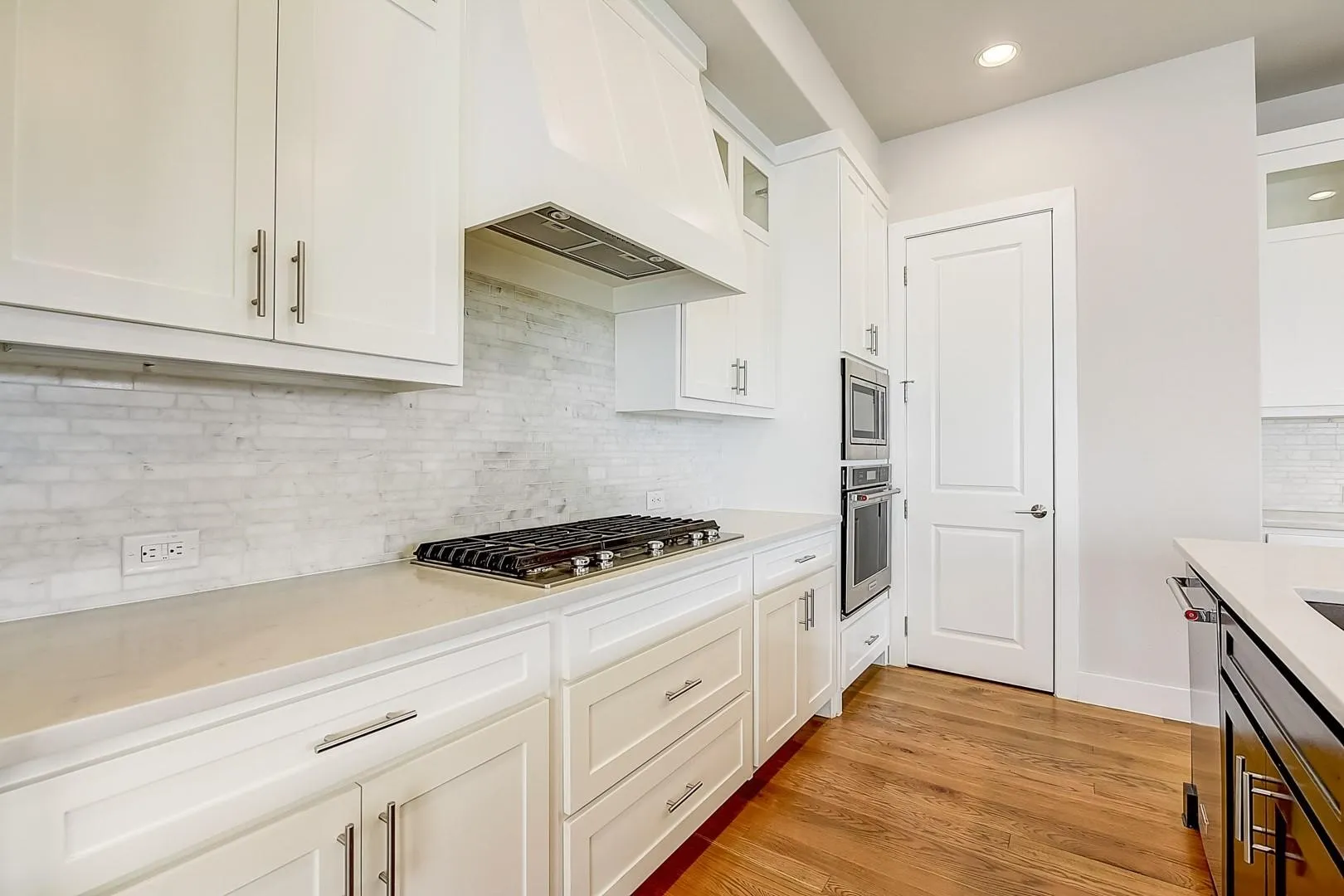 White cabinetry with brushed nickel hardware, stainless steel gas range, built-in double oven, light-toned countertops, and wood-finish flooring