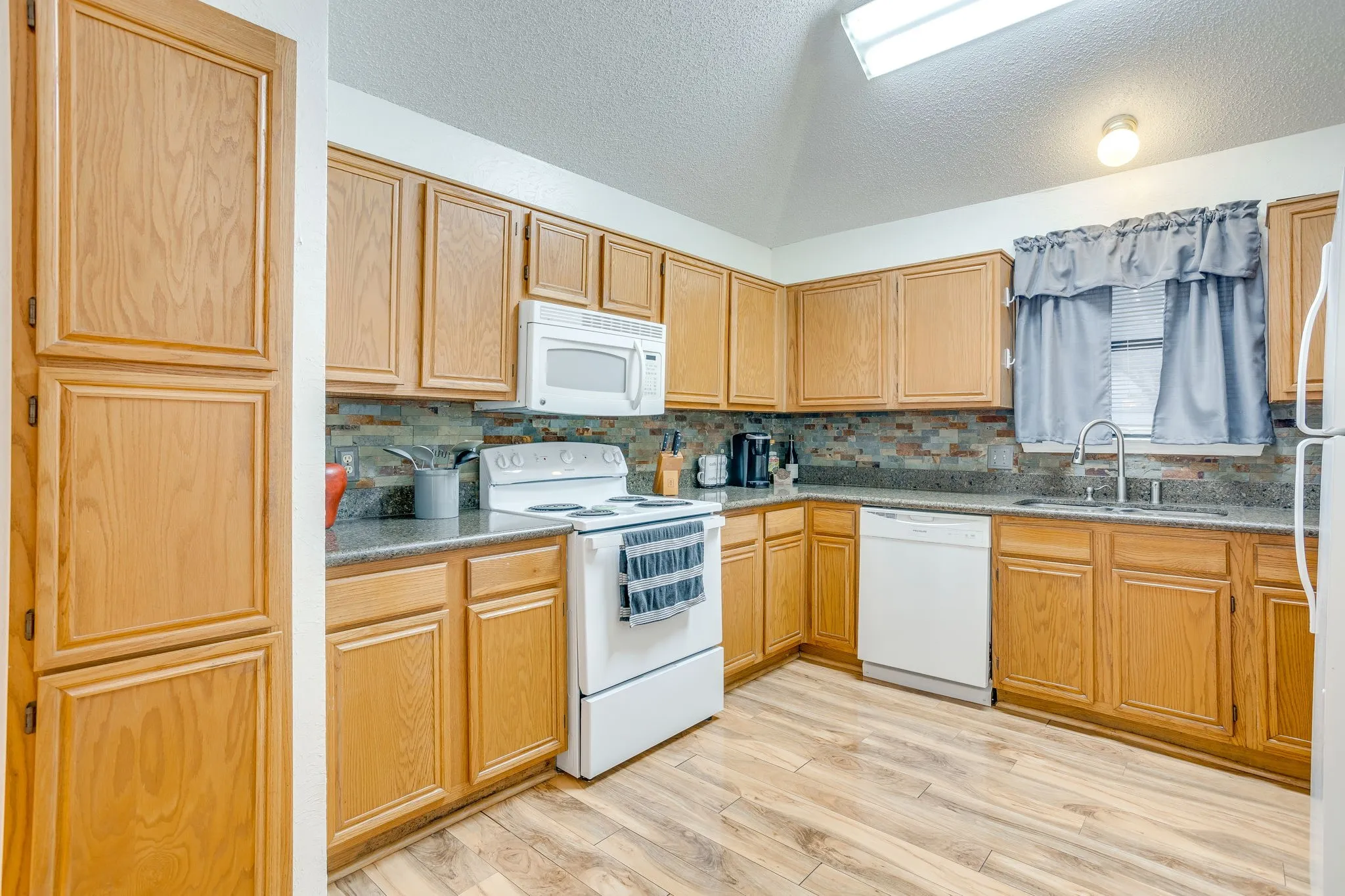 Spacious kitchen featuring warm wood cabinetry, white appliances, tile countertops with coordinating backsplash, a window overlooking the exterior, and wood-finish flooring throughout the cooking and dining area