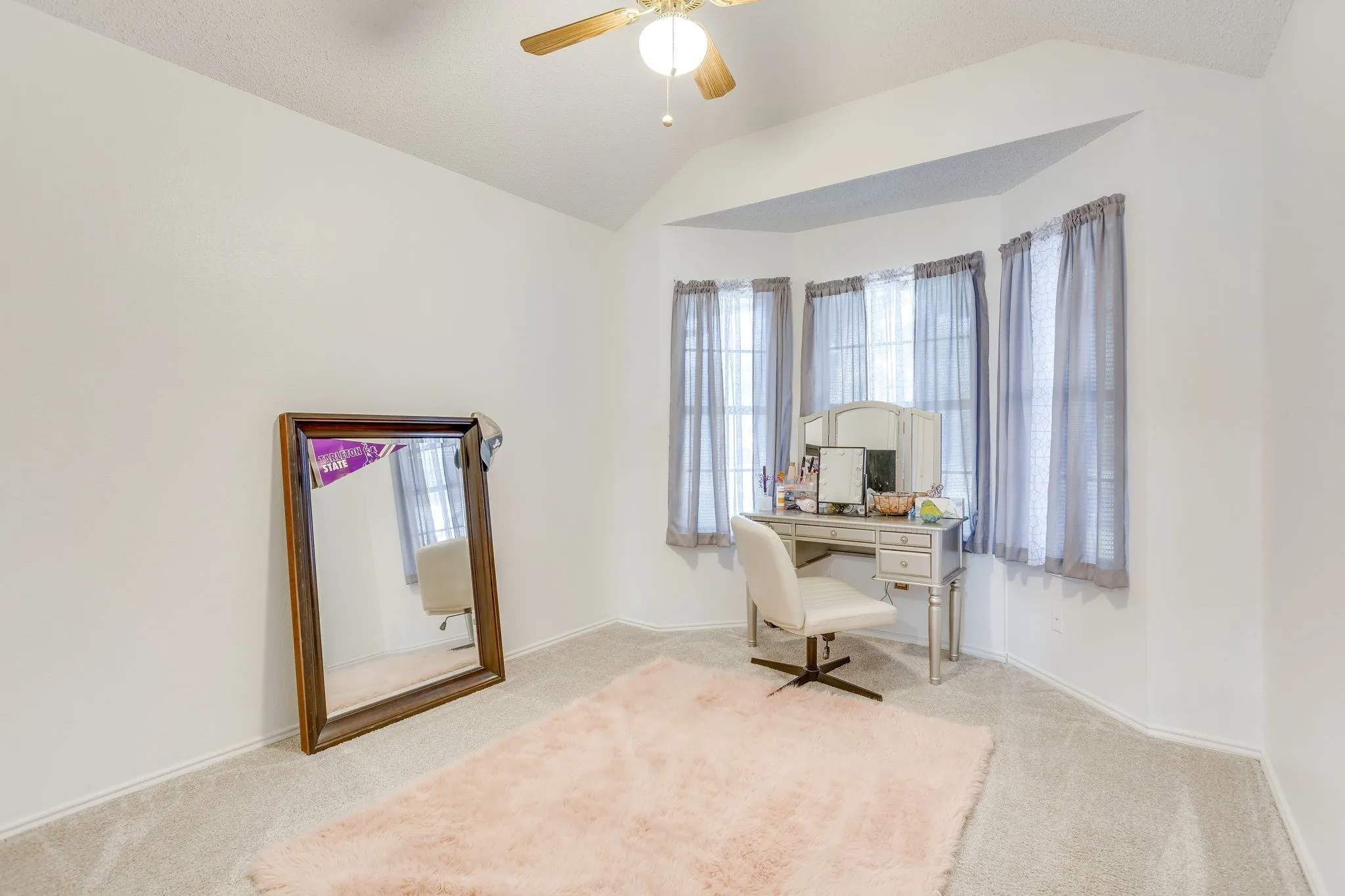 Bright secondary bedroom offering abundant natural light from multiple windows with soft blue drapery, neutral carpet flooring, and a ceiling fan with integrated lighting